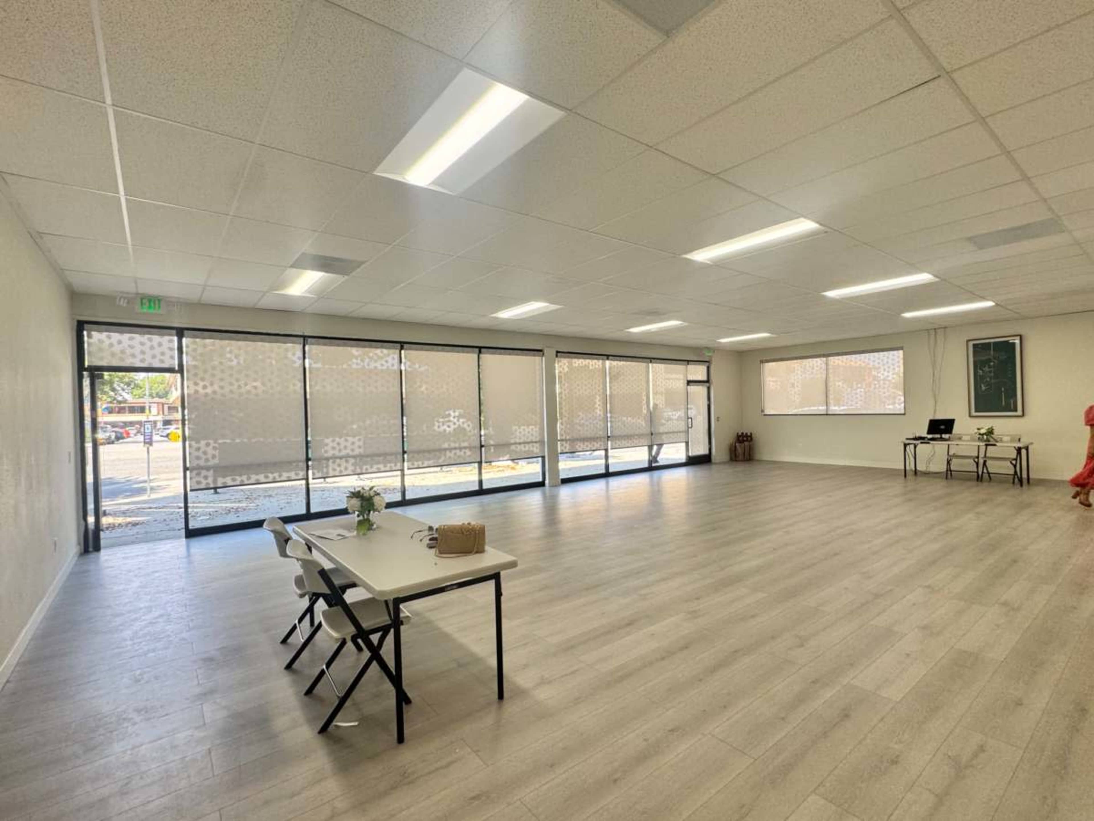 The image shows a spacious, empty room with large windows and light-colored flooring, featuring two tables and chairs arranged on one side.