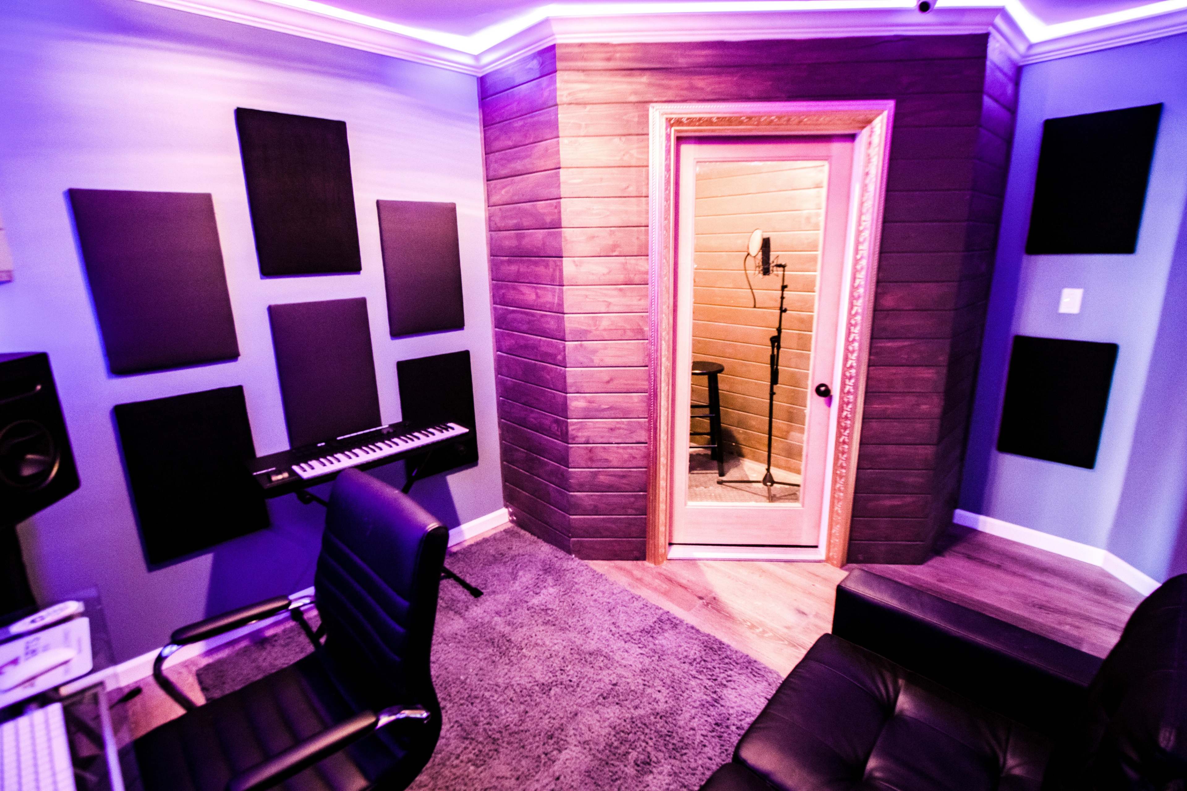 A modern recording studio with soundproof panels on the walls, a keyboard, and a vocal booth visible through a door.