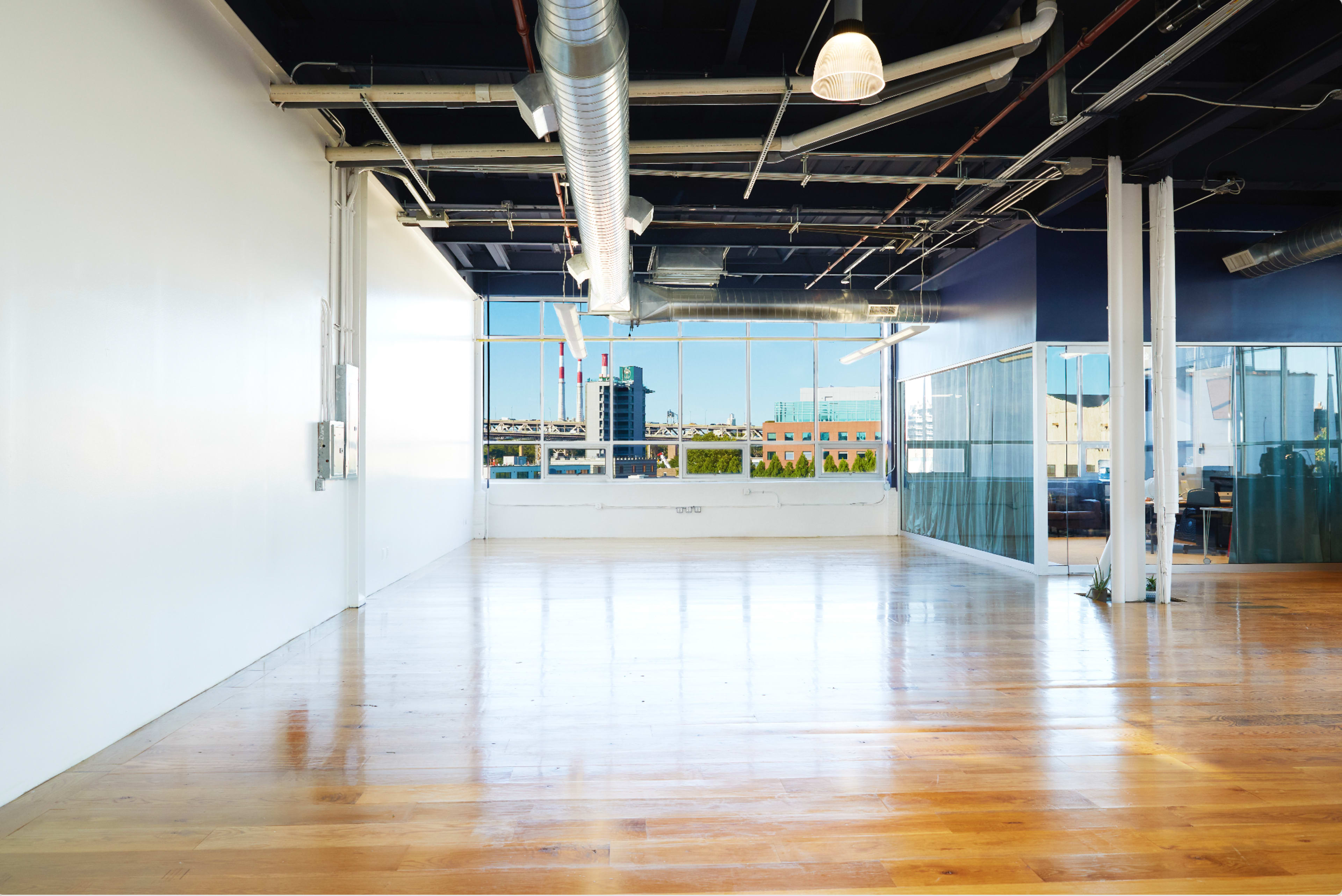The image shows a spacious, empty office interior with wooden flooring, large windows, and industrial-style ceiling fixtures.