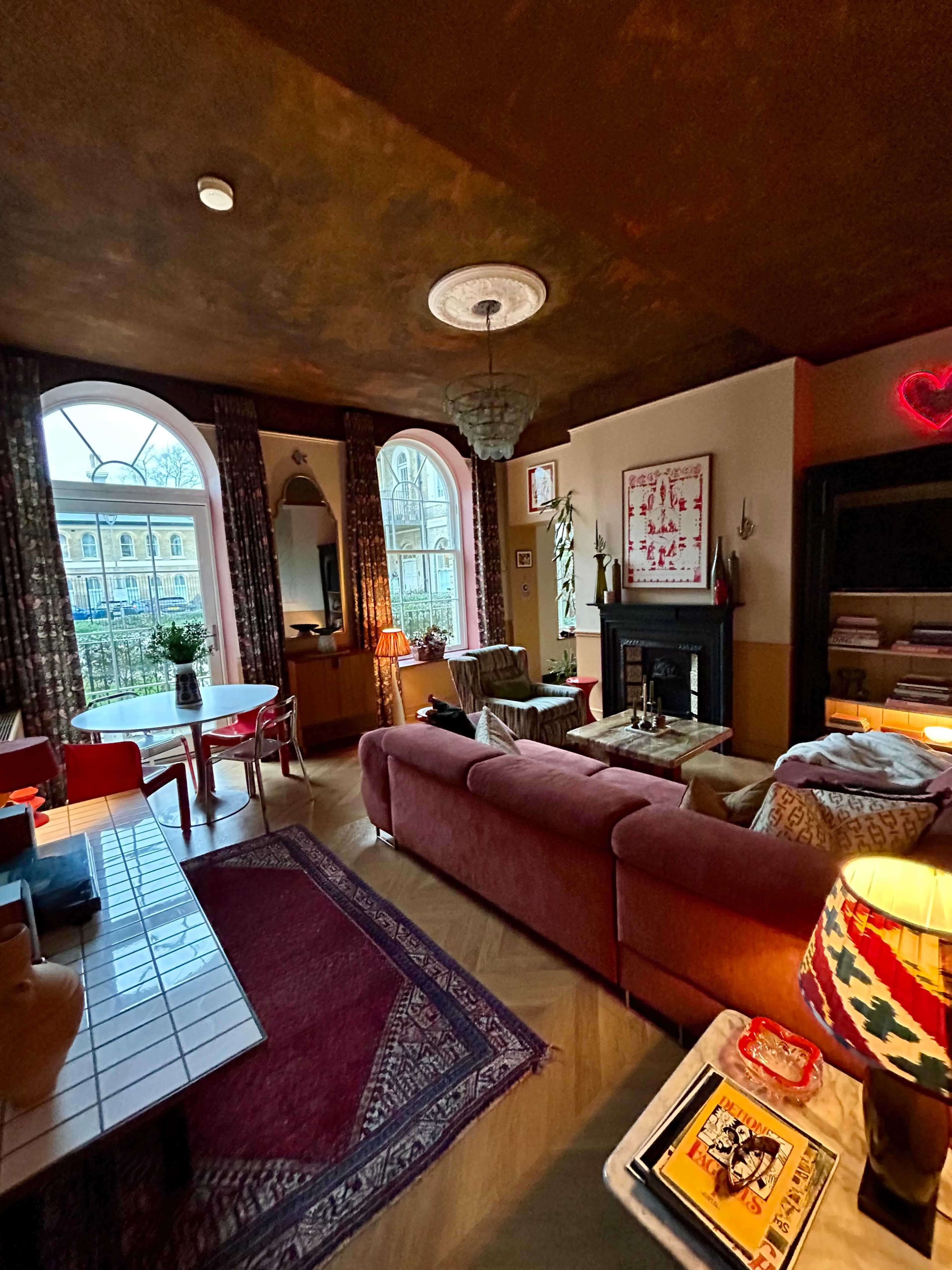 The image shows a cozy living room featuring a pink sofa, a wooden table with red chairs, a fireplace, and large windows allowing natural light to enter.