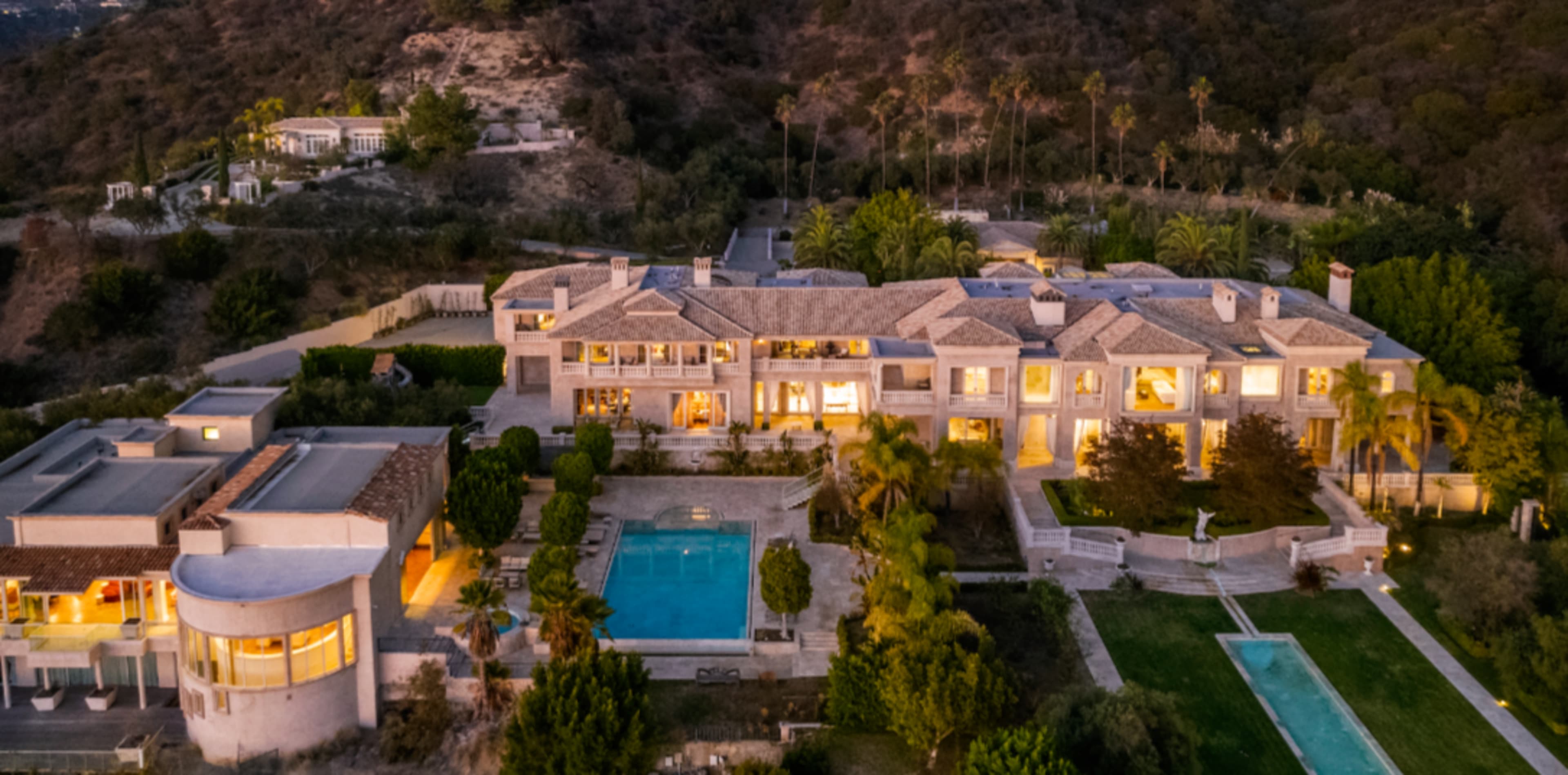 The image shows a large, luxurious house with multiple stories, a swimming pool, and landscaped gardens situated on a hillside.