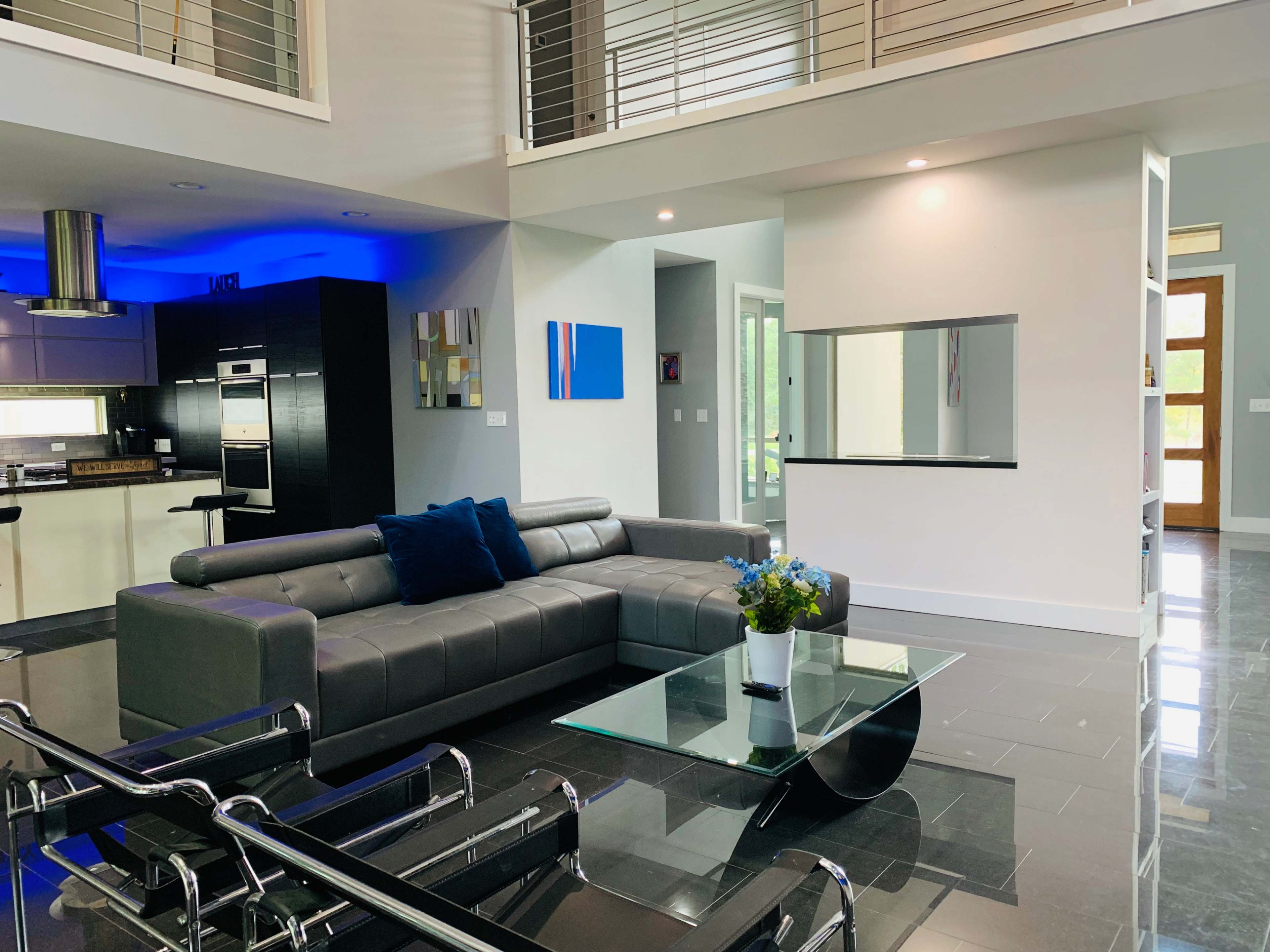 The image shows a modern living room with a gray sectional sofa, a glass coffee table, and an open layout leading to a contemporary kitchen with blue lighting.