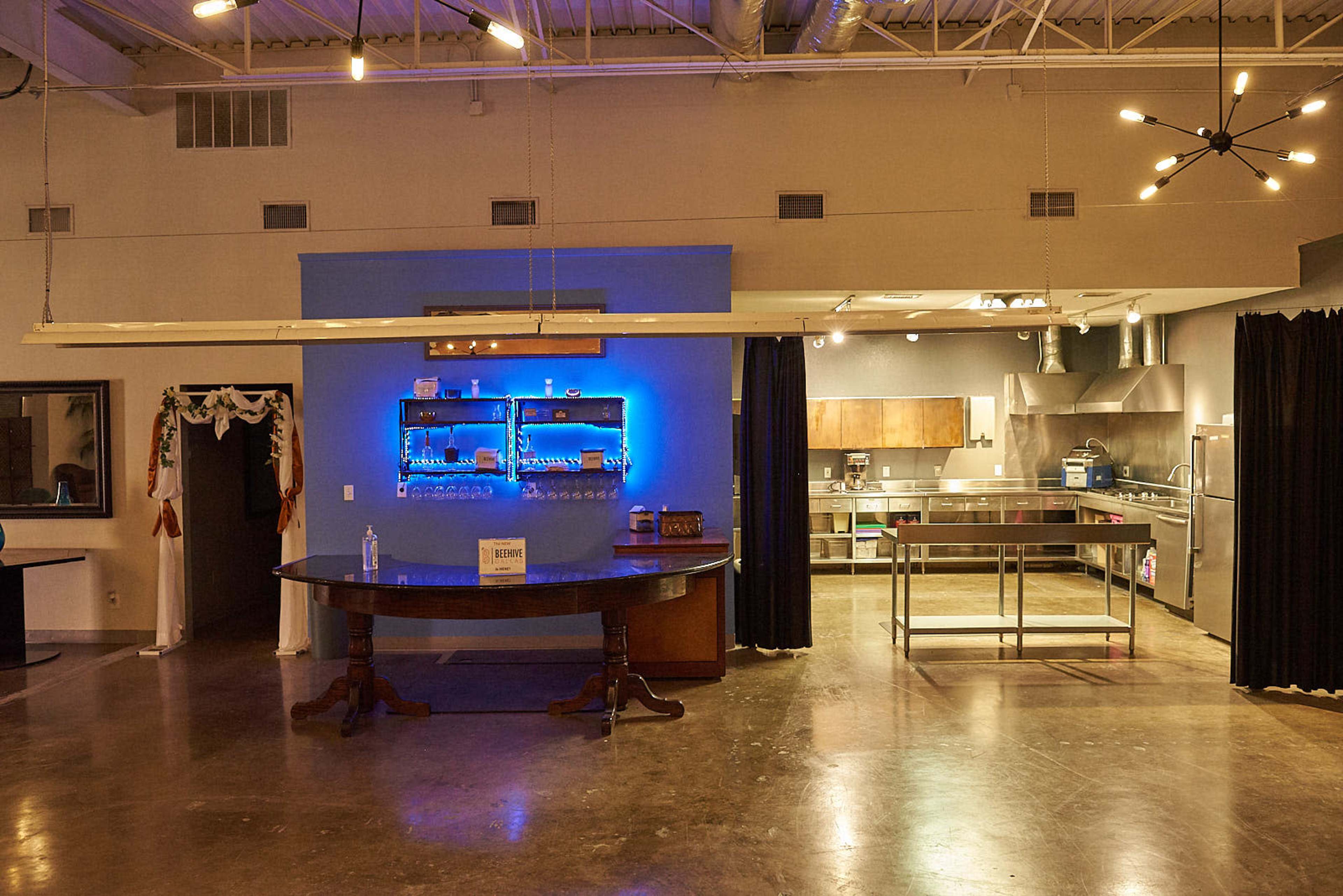 An industrial-style kitchen and dining area featuring a large round table, illuminated shelves with blue lighting, and stainless-steel kitchen equipment.