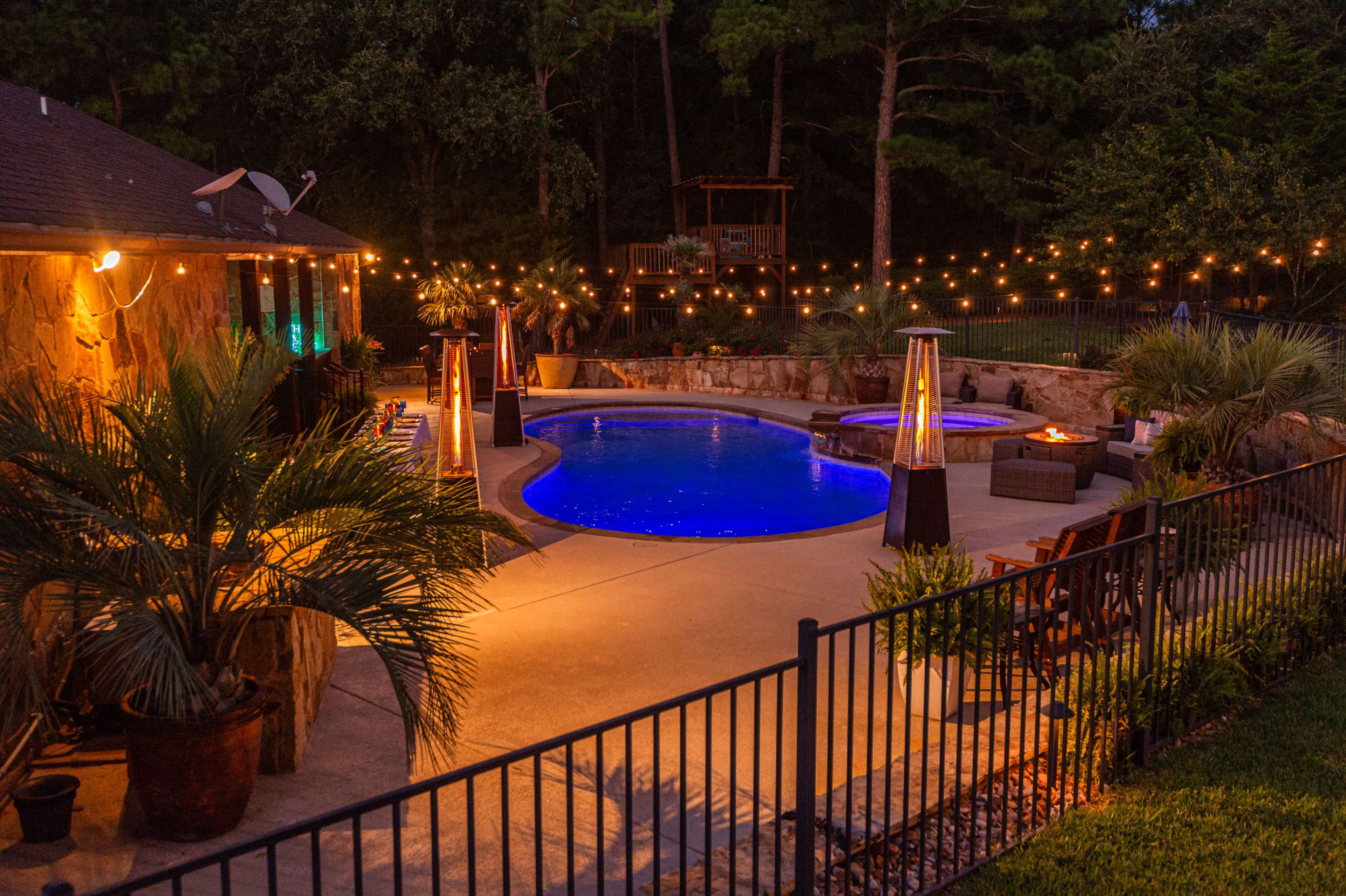 A pool area featuring illuminated water and surrounding seating is adorned with string lights and a fire pit, set against a backdrop of trees at night.