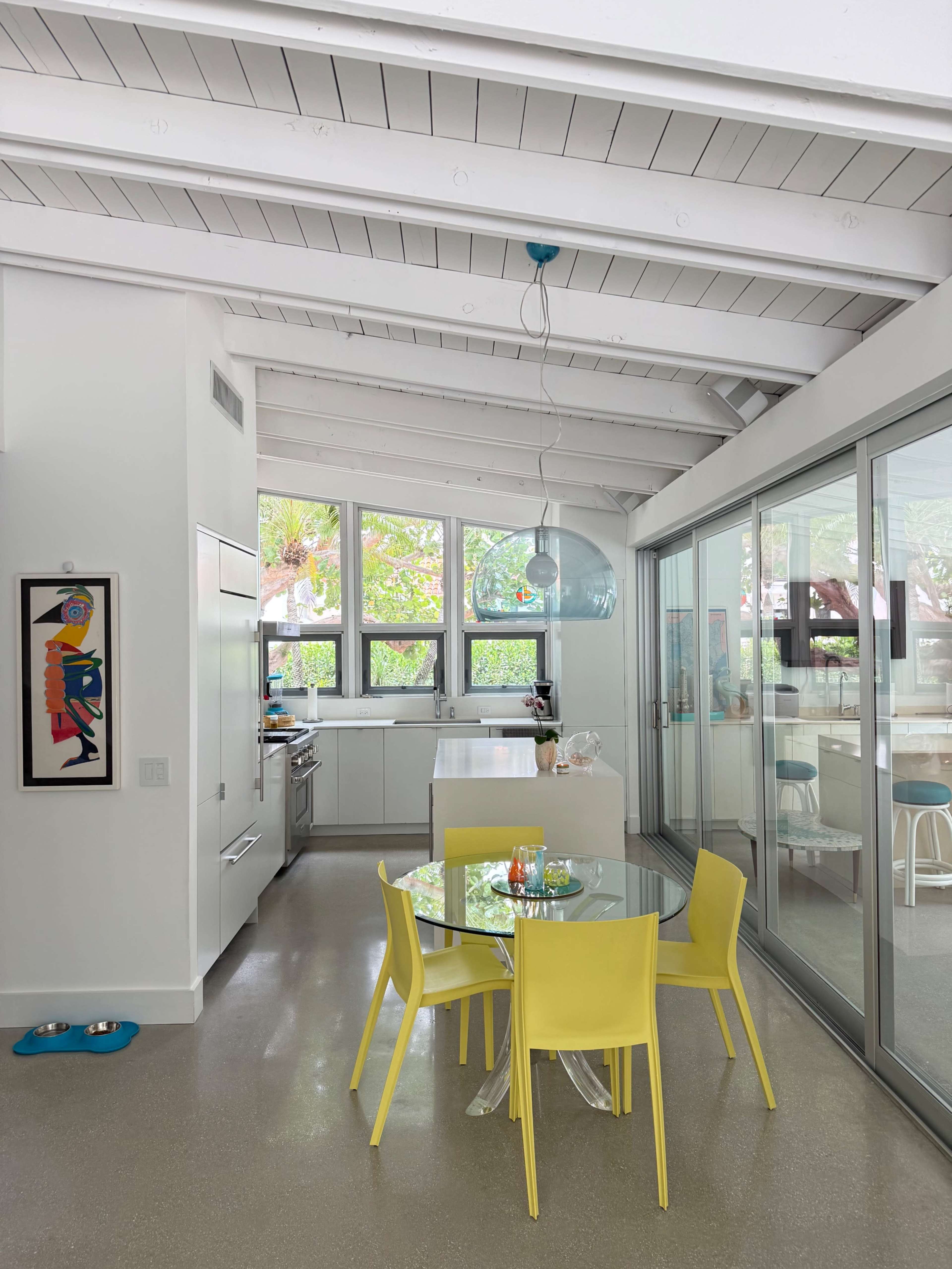 The image shows a modern kitchen and dining area with a round glass table surrounded by bright yellow chairs, large windows letting in natural light, and an open layout.