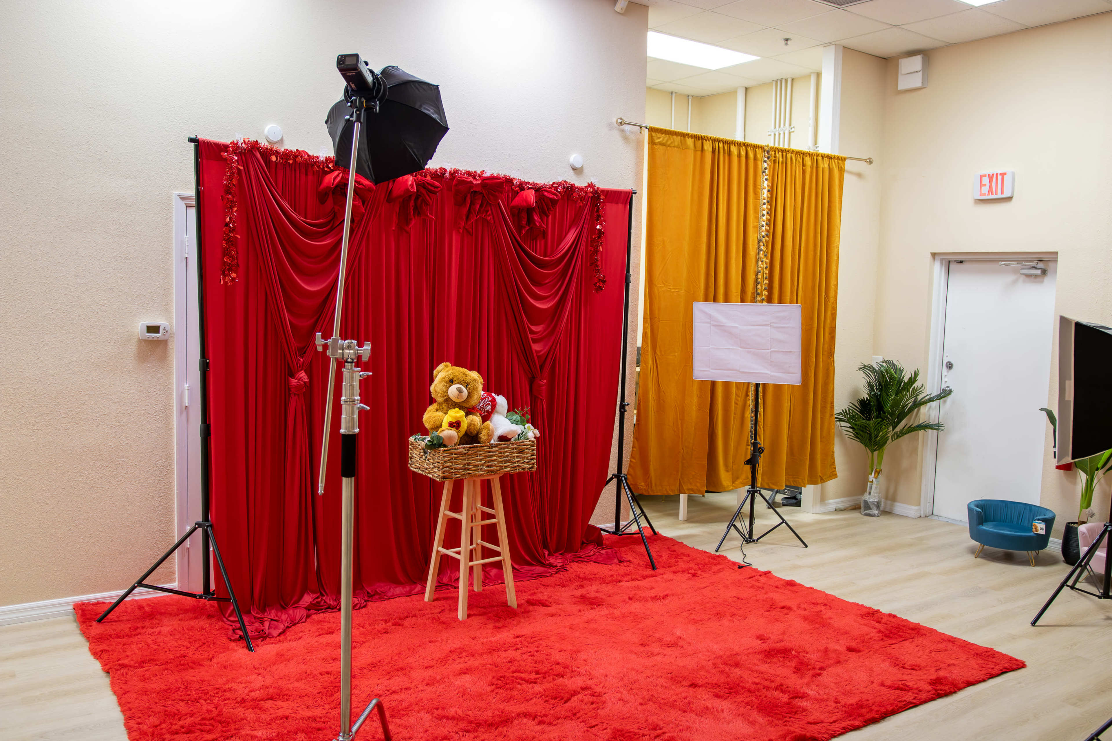 The image shows a photography studio setup with a red backdrop, a stool with stuffed animals, and various lighting equipment.