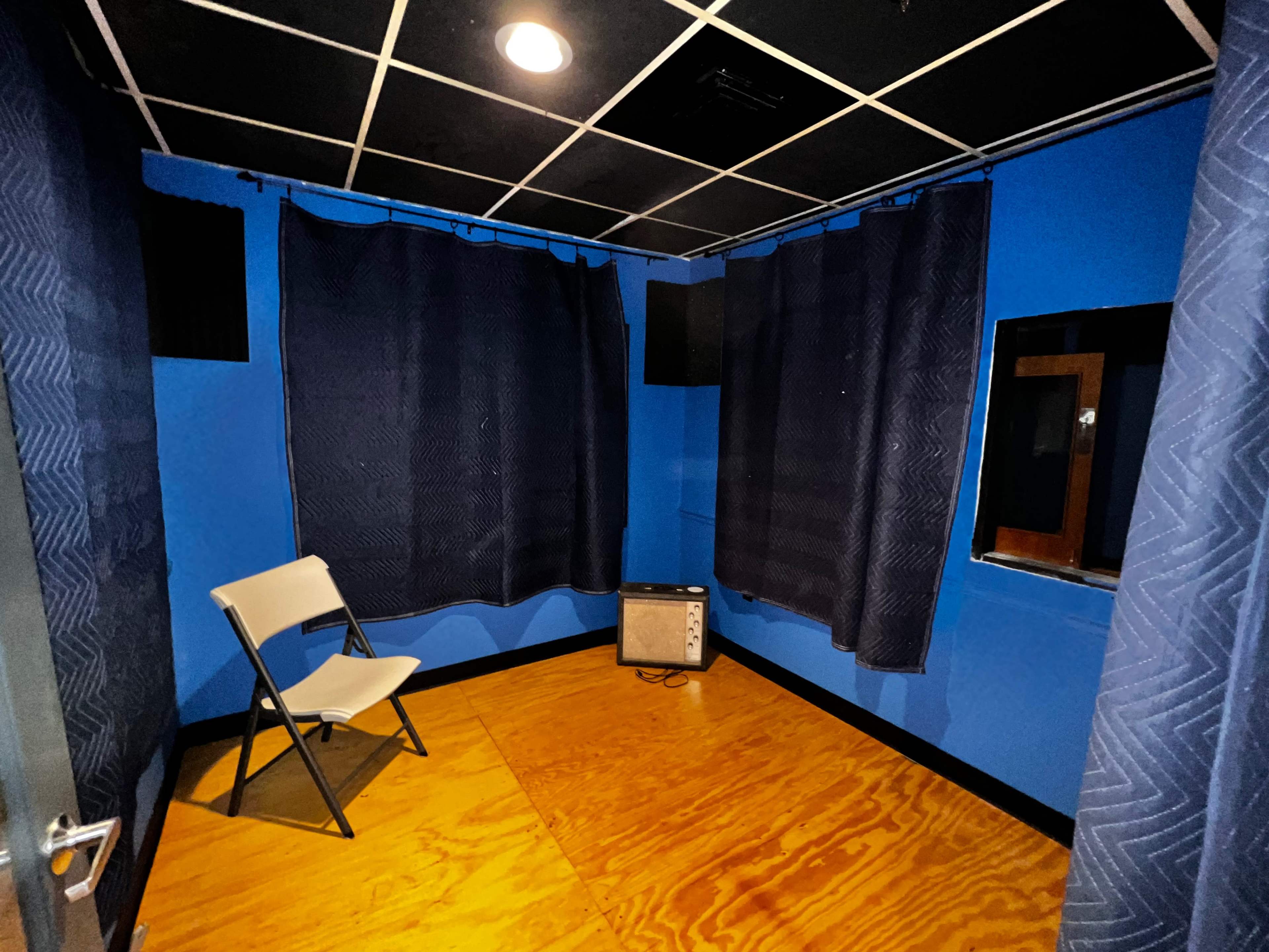 The image shows a small blue room with a folding chair, curtains, and an amplifier on the floor.