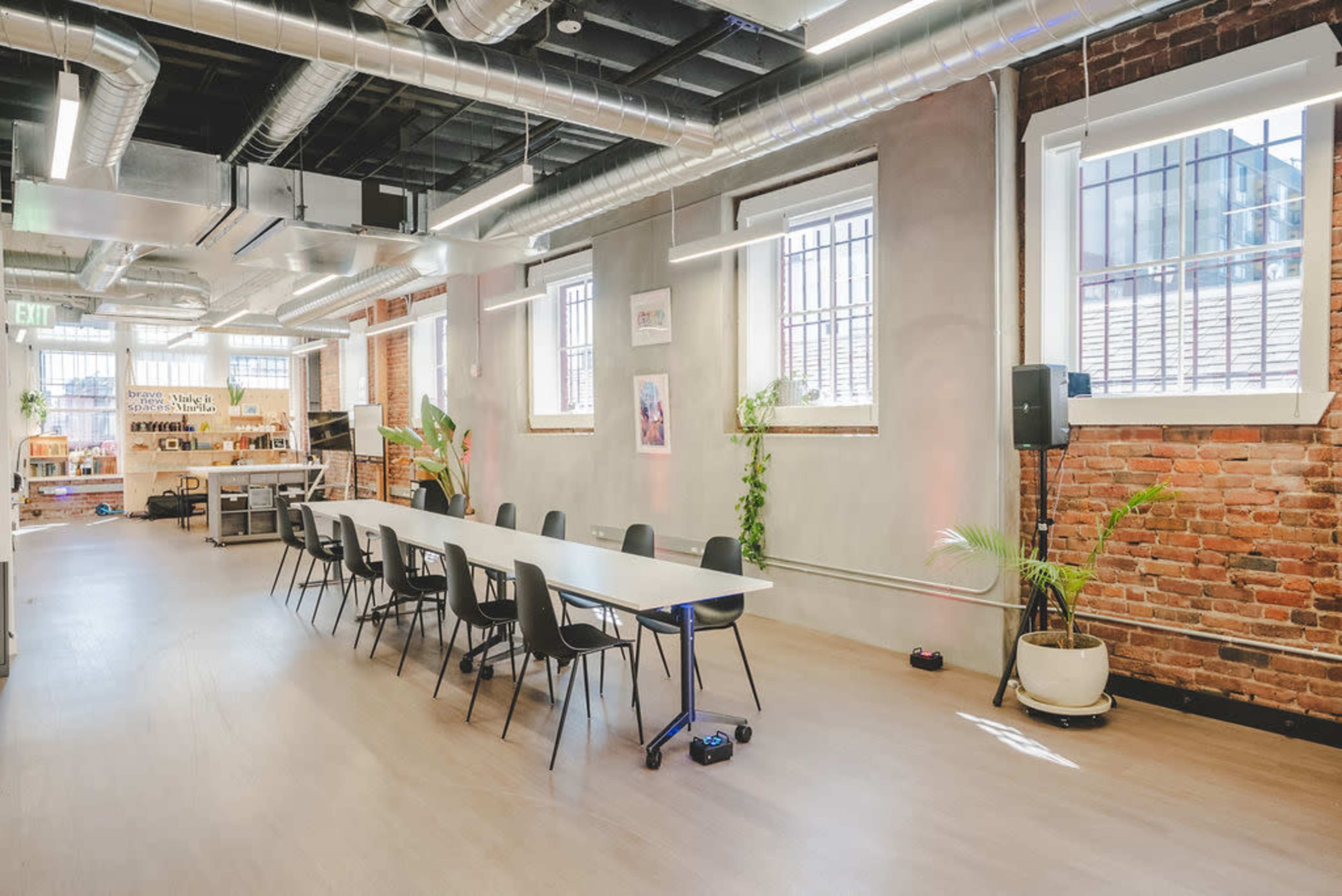 The image shows a modern, open-plan office space featuring a long conference table surrounded by black chairs, with exposed brick walls and large windows letting in natural light.