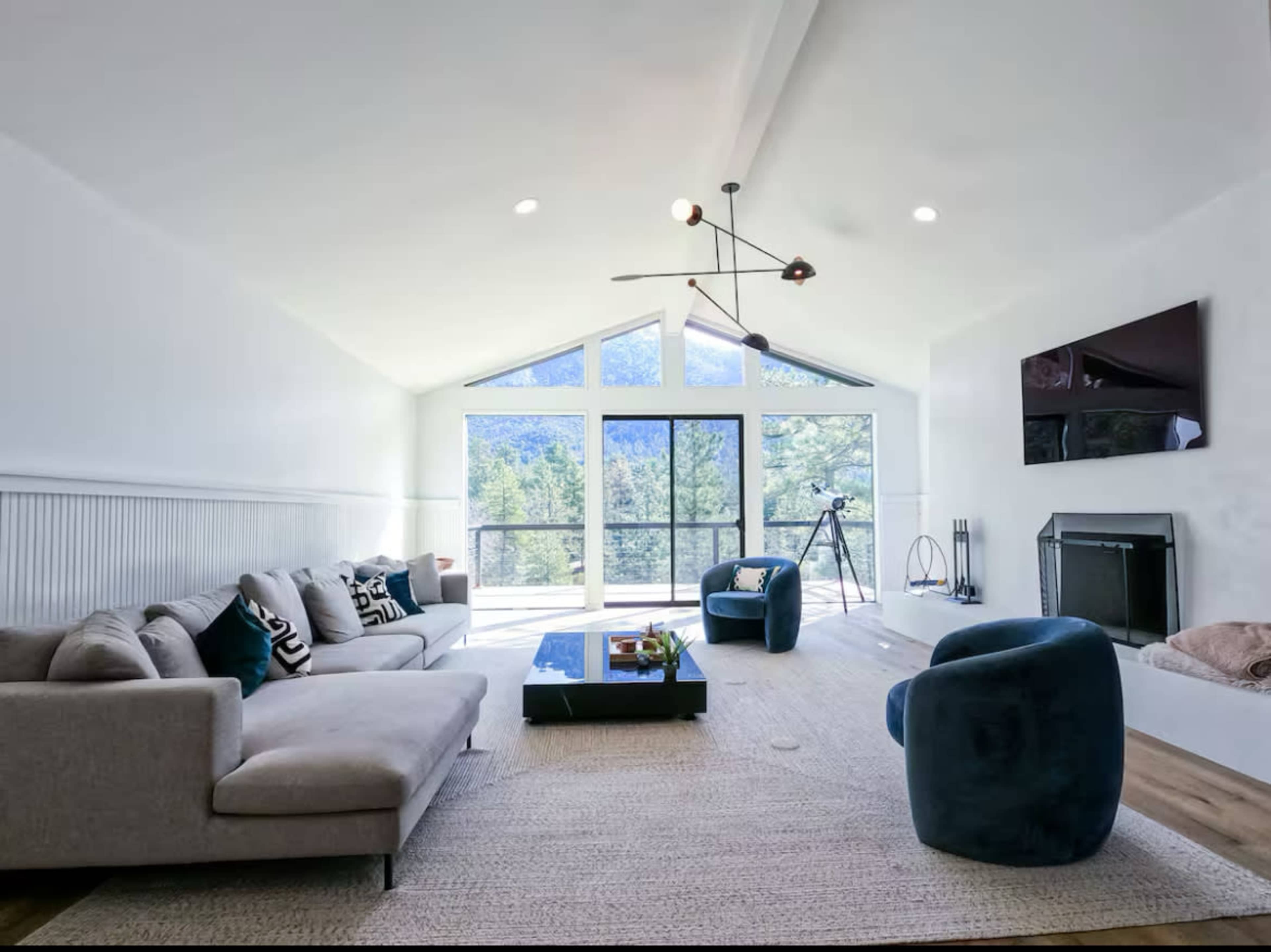 The living room features a modern design with large windows overlooking a scenic view, a light-colored sectional sofa, two blue accent chairs, and a fireplace.