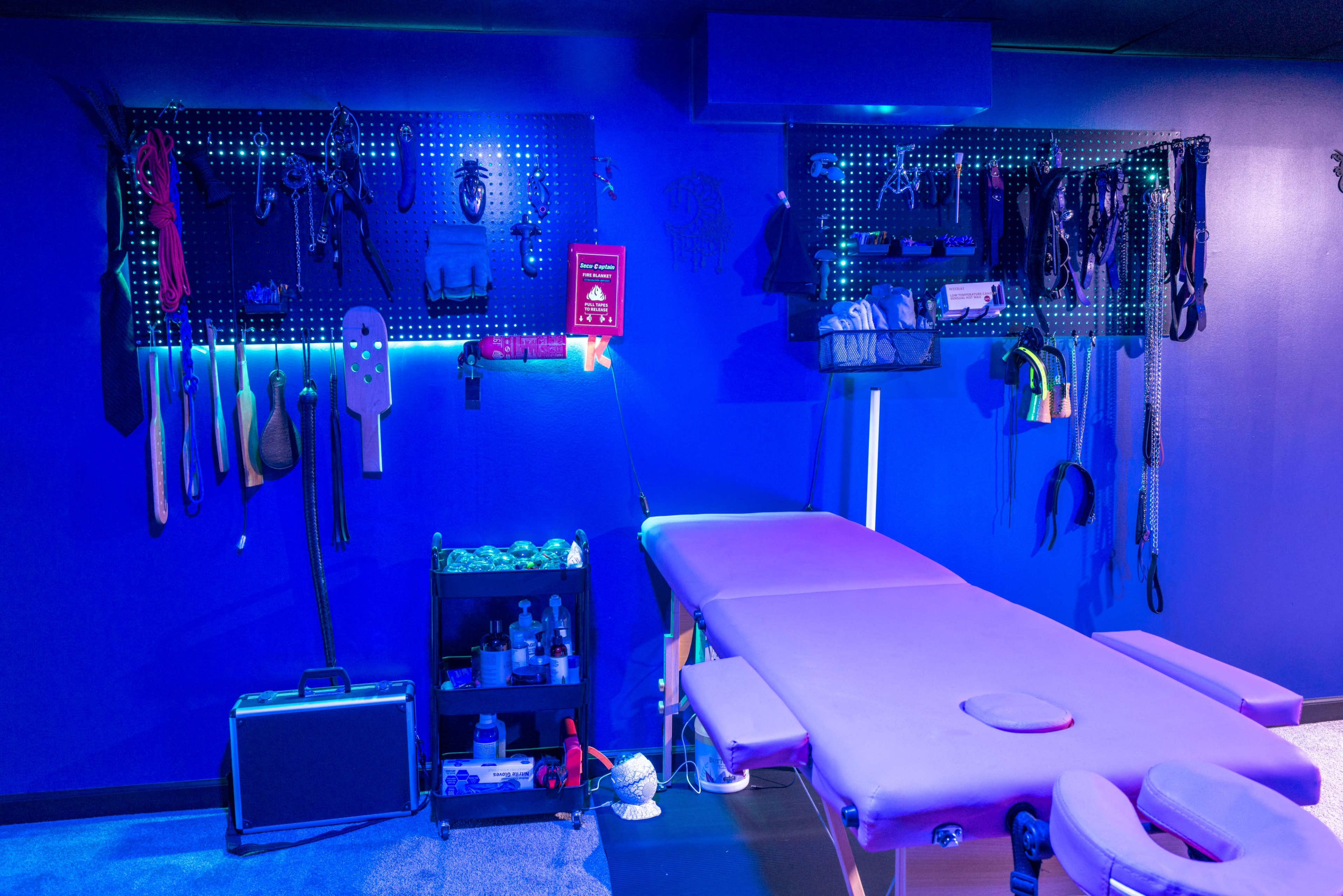 The image features a dimly lit massage room with a padded table and various tools and equipment hanging on the walls.
