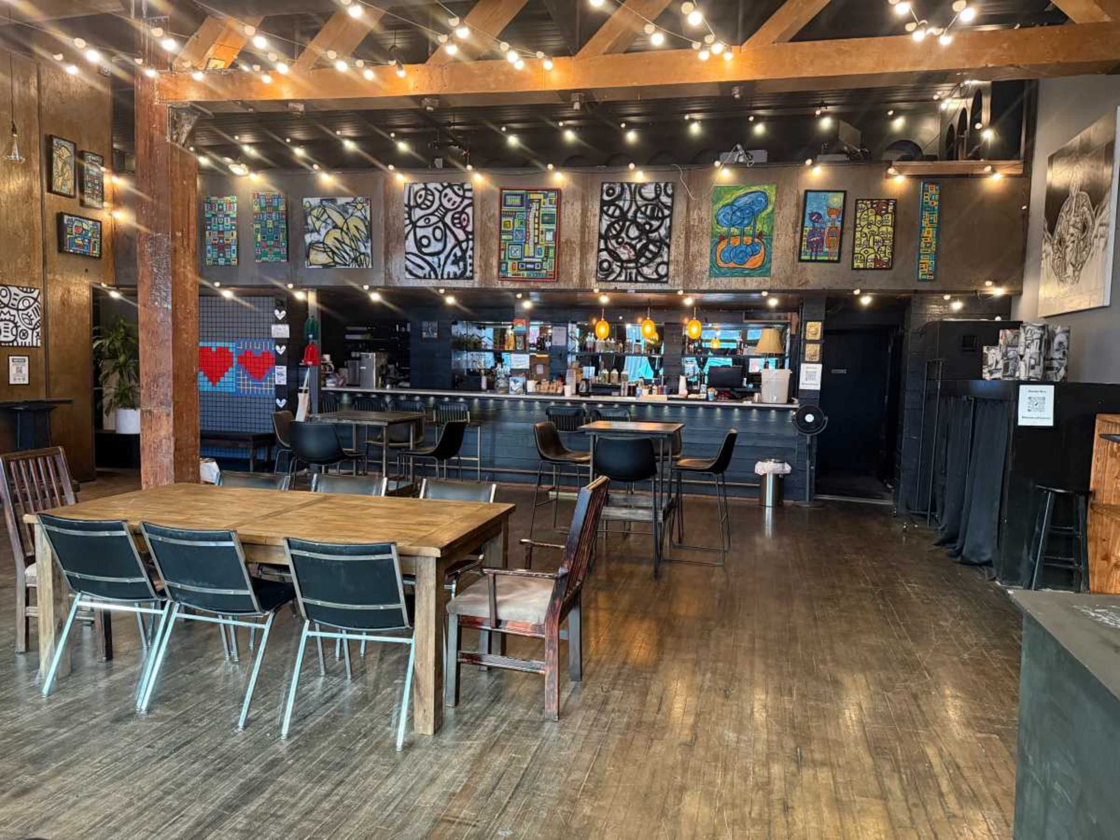 The image shows a spacious café interior featuring a wooden table with chairs, a bar area with colorful artwork on the walls, and string lights hanging from the ceiling.