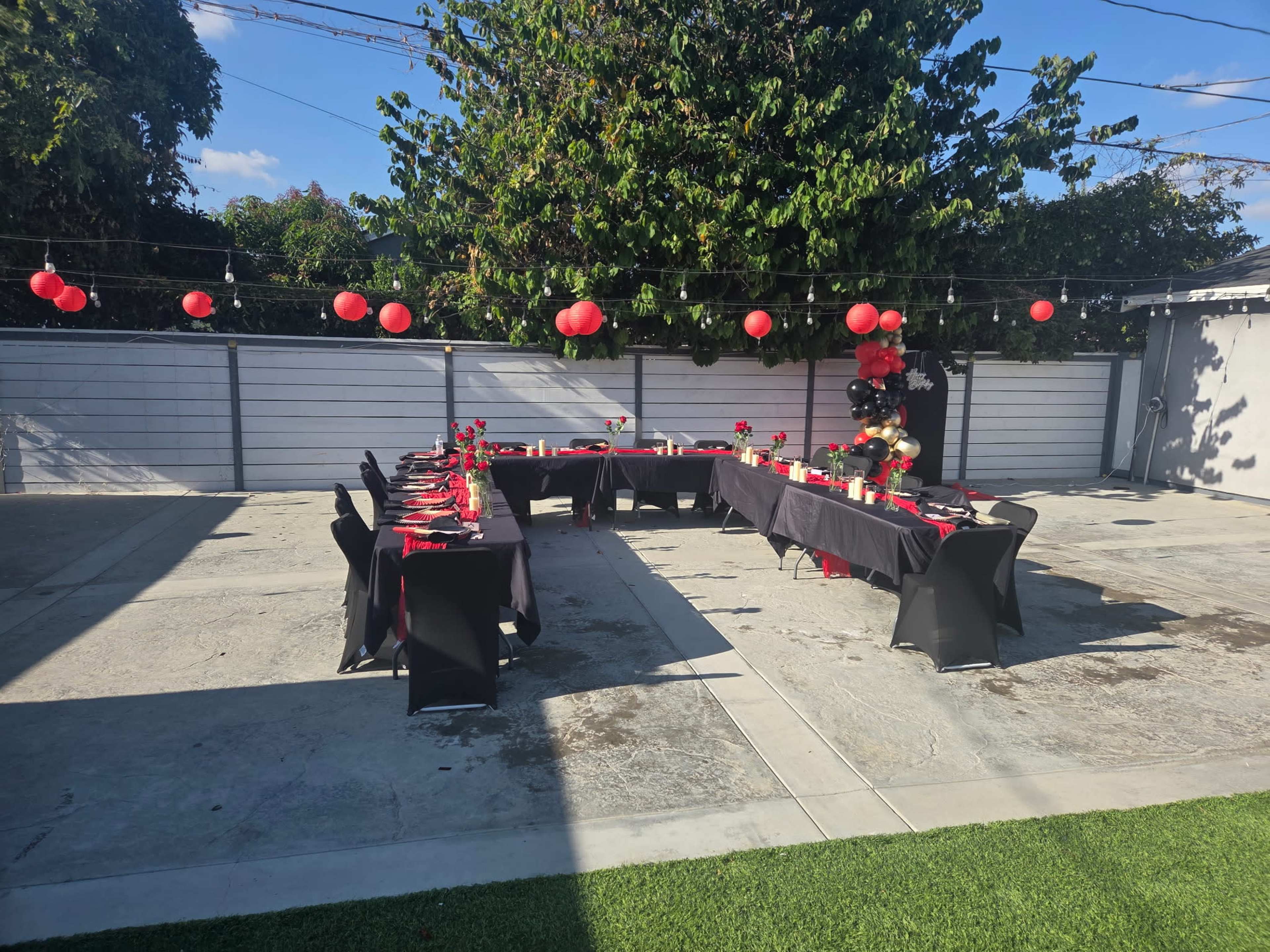 A spacious outdoor area features two long tables set for an event, adorned with black and red decorations, surrounded by colorful lanterns and greenery.