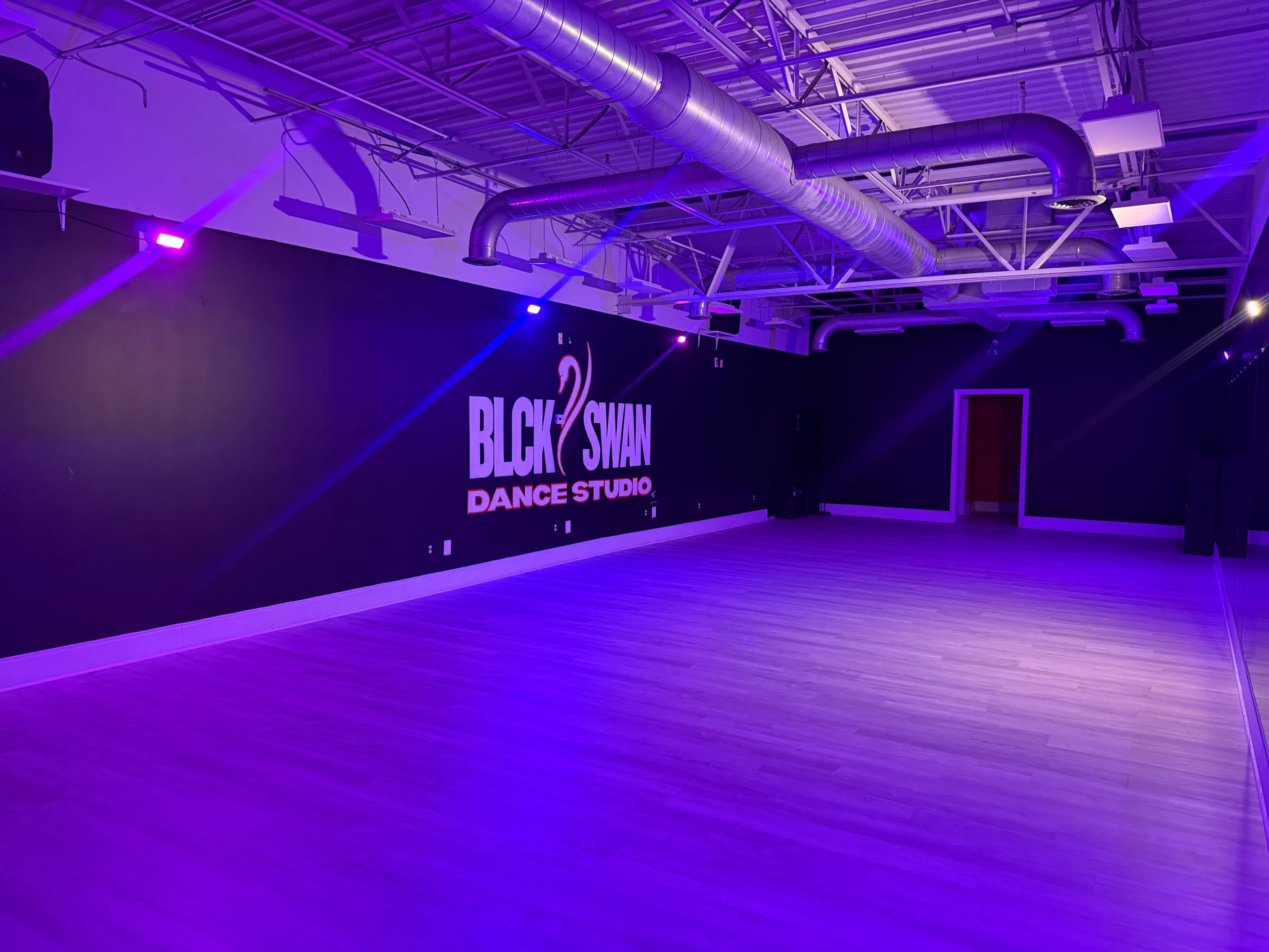 The image shows an empty dance studio with purple lighting and a wall featuring the logo "BLCK SWAN DANCE STUDIO."