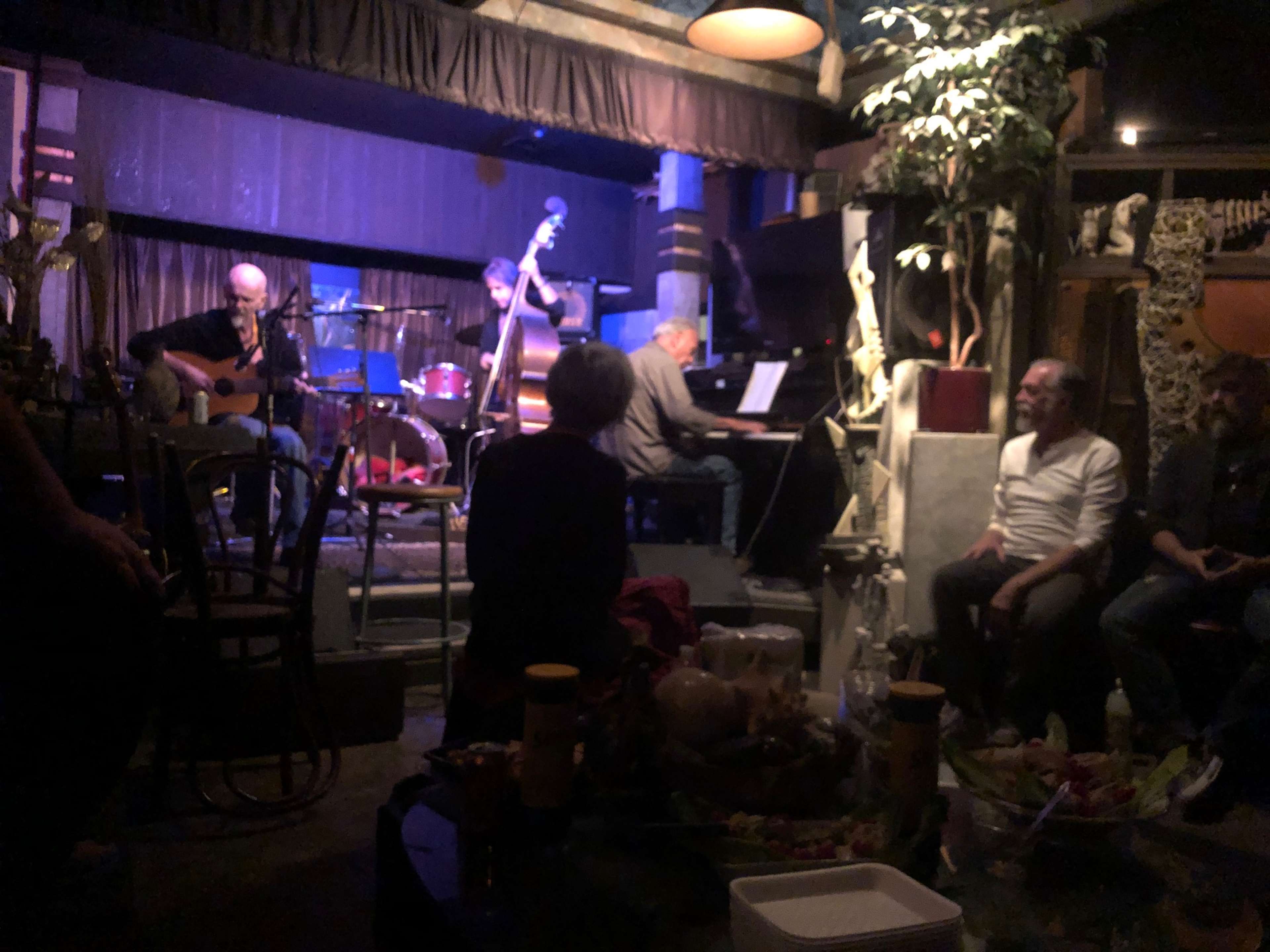 A small jazz band performs on a dimly lit stage in a cozy venue while an audience sits nearby.