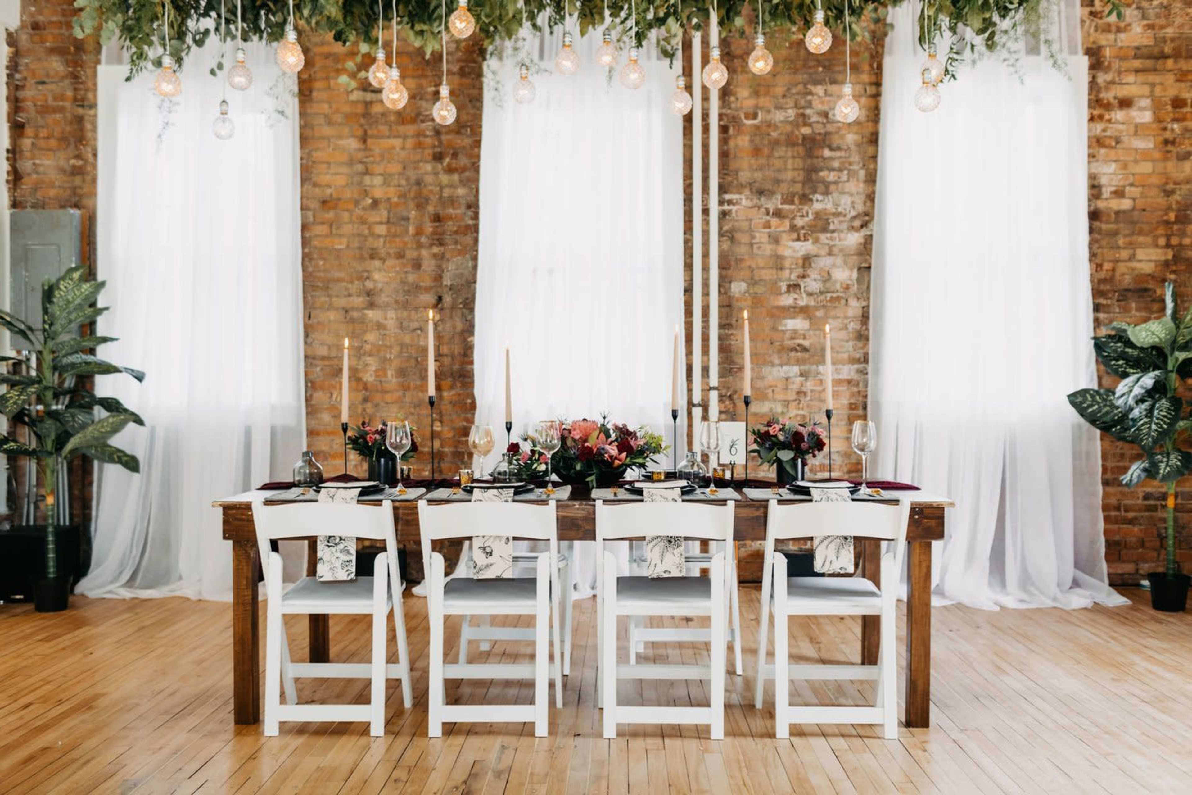 A long wooden dining table is set with elegant dinnerware and surrounded by white chairs under hanging light fixtures and draped fabric in a rustic setting.