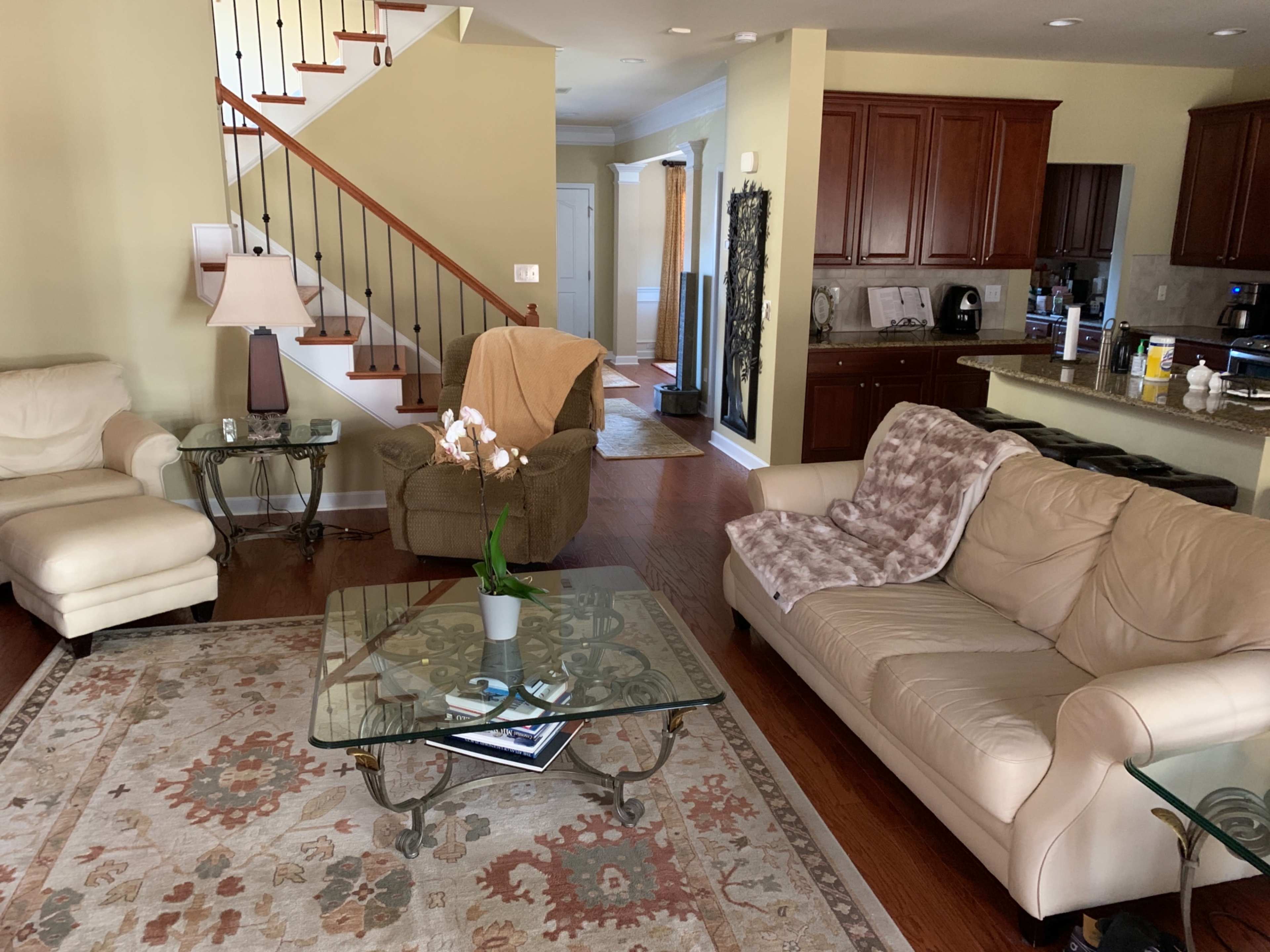 A living room features two cream-colored sofas, a glass coffee table, and a staircase in the background, with a dining area visible through an open doorway.