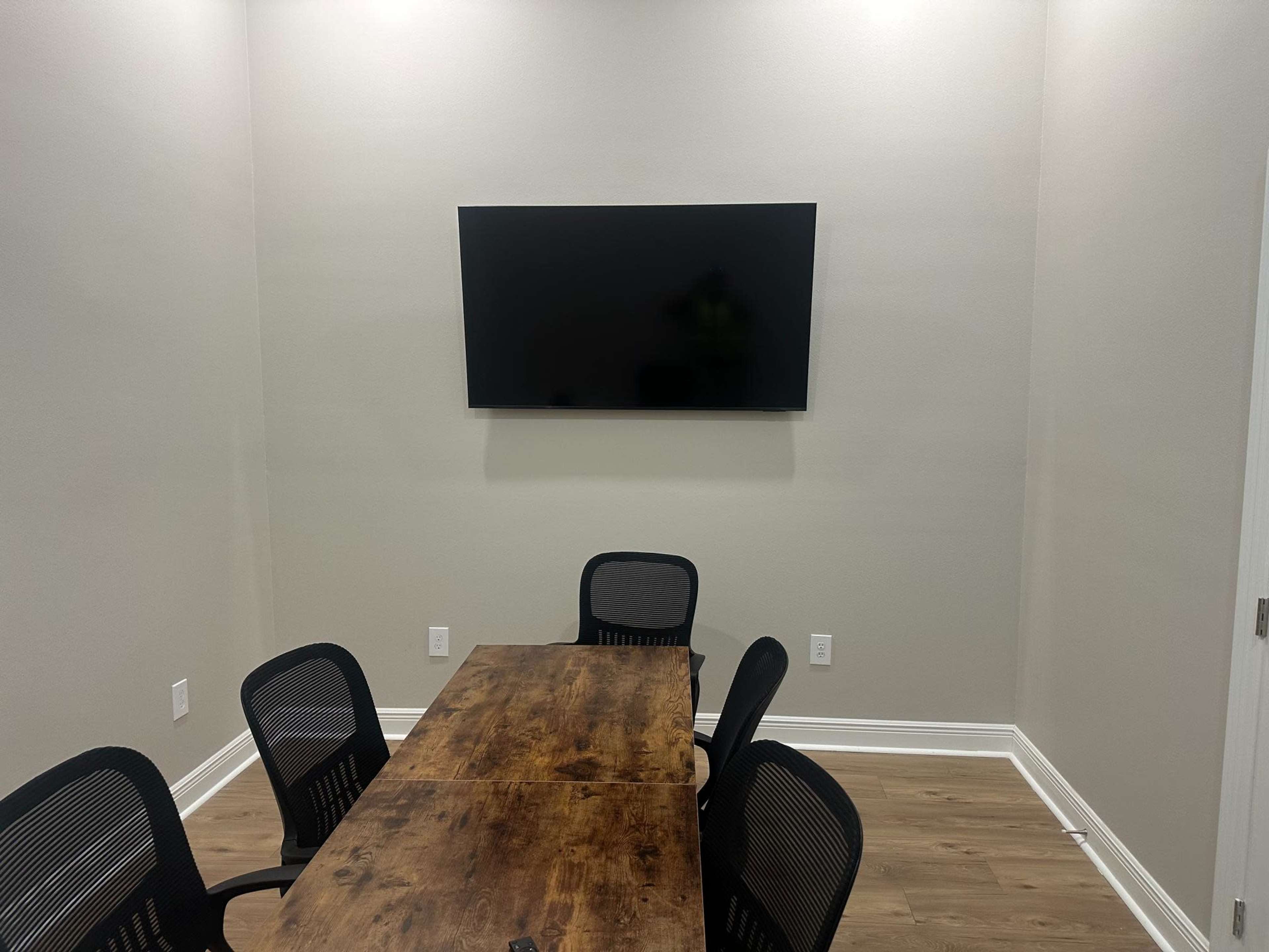 A conference room features a rectangular wooden table surrounded by black mesh chairs, with a large flat-screen monitor mounted on the wall.