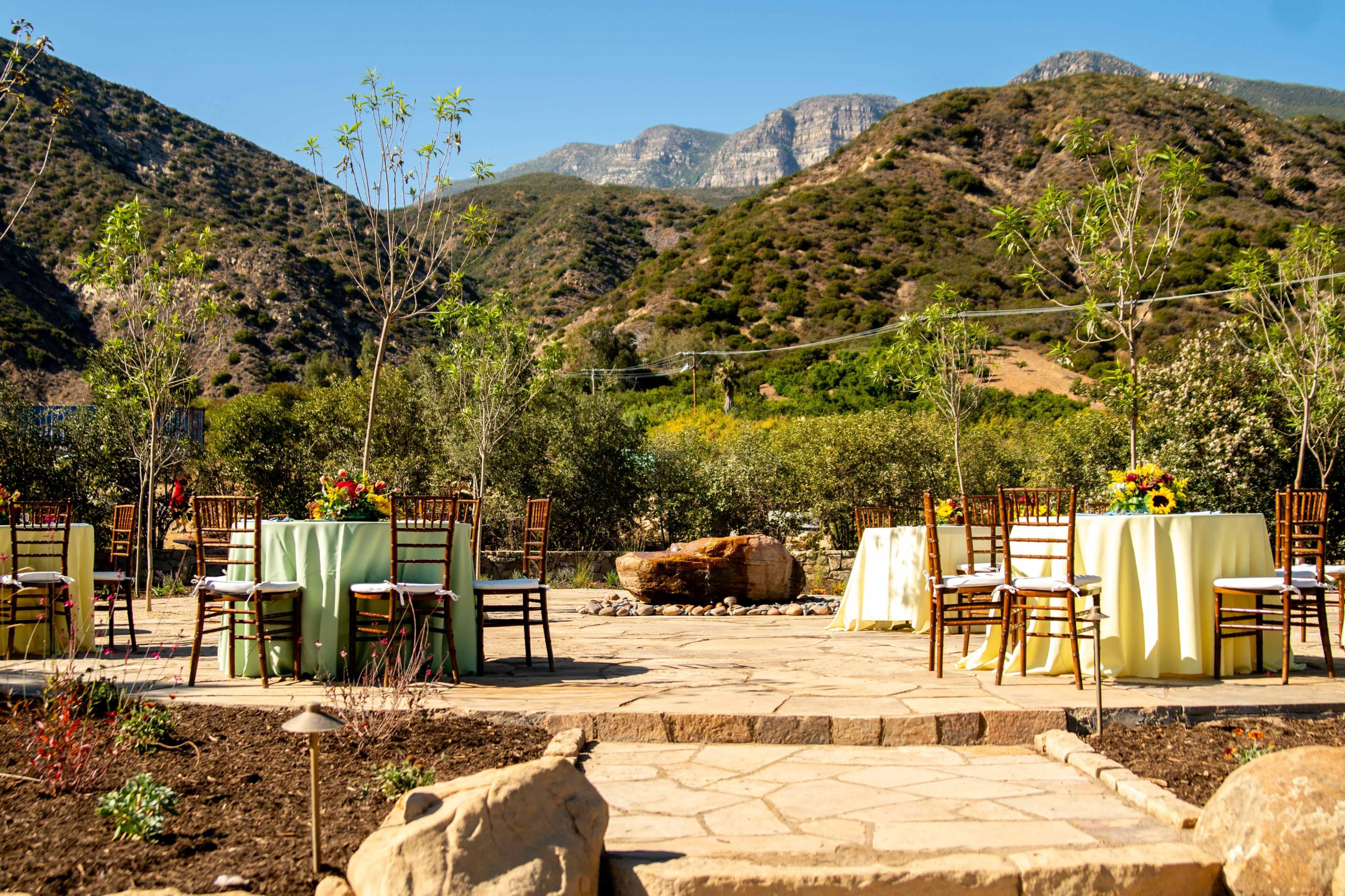 An outdoor event space features tables with yellow linens set up against a backdrop of mountains and greenery.