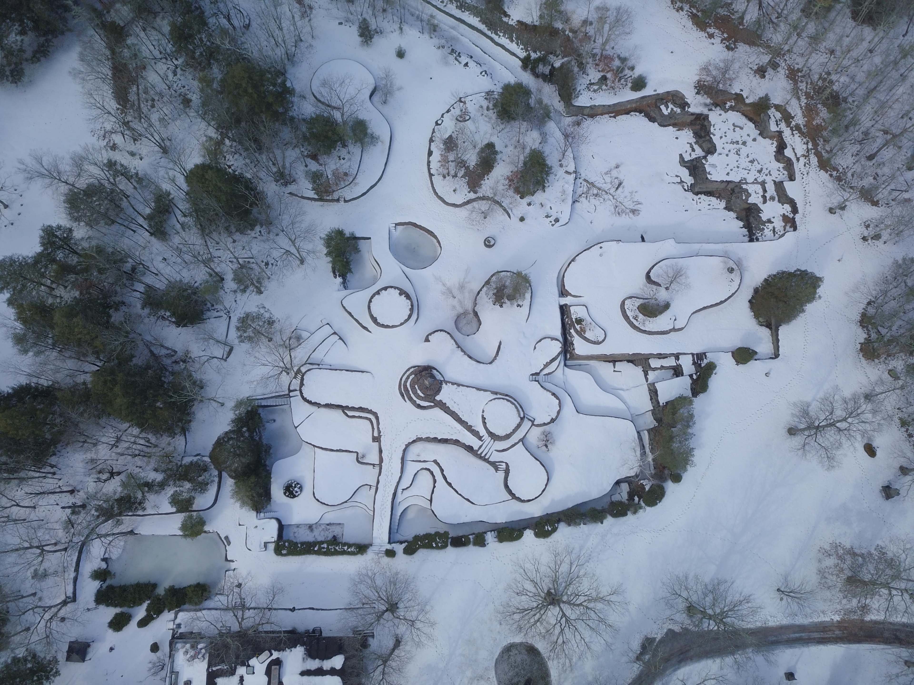An aerial view shows a snow-covered landscape featuring winding paths, garden beds, and a cluster of houses.