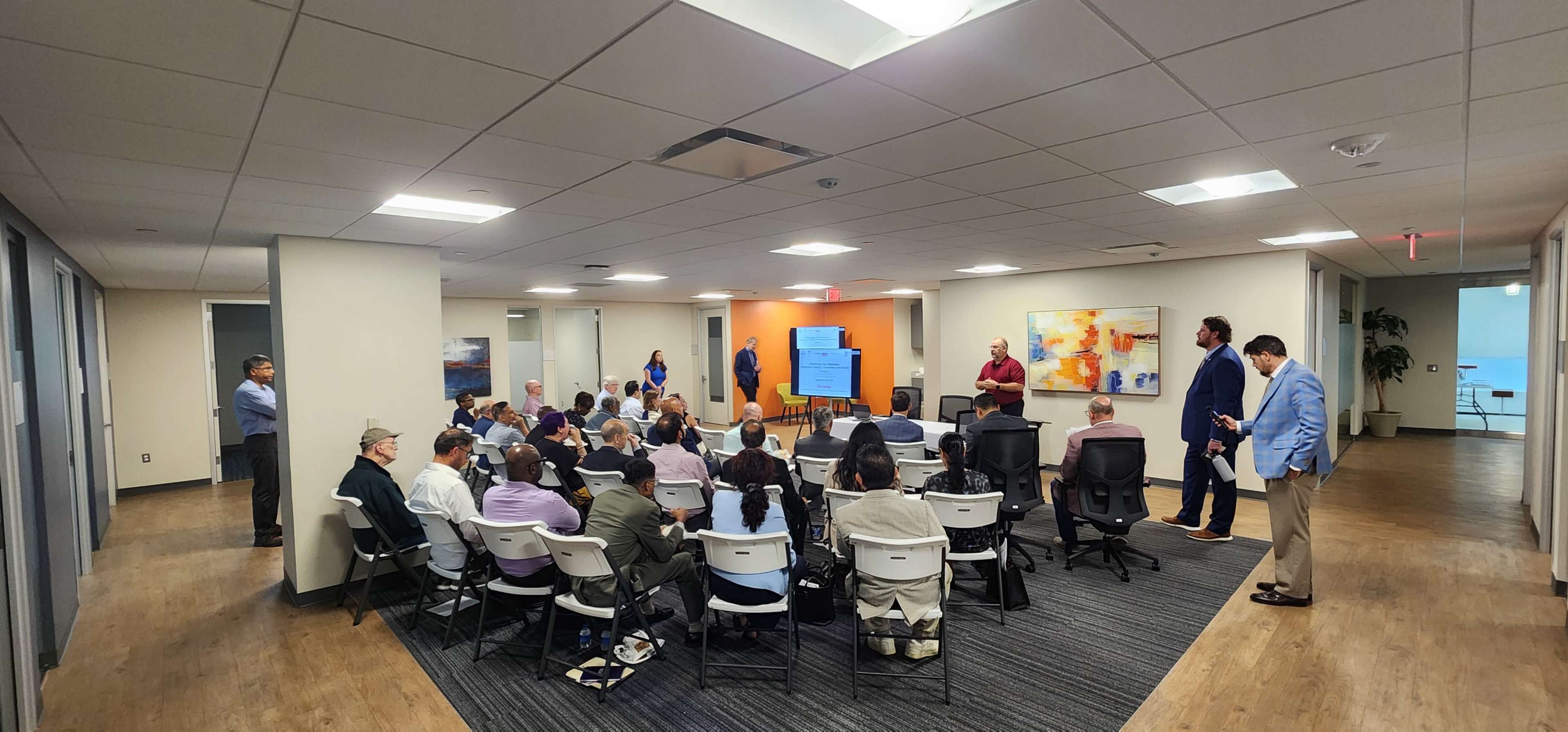 A diverse group of people is seated in a conference room while others engage with a presentation on a screen.