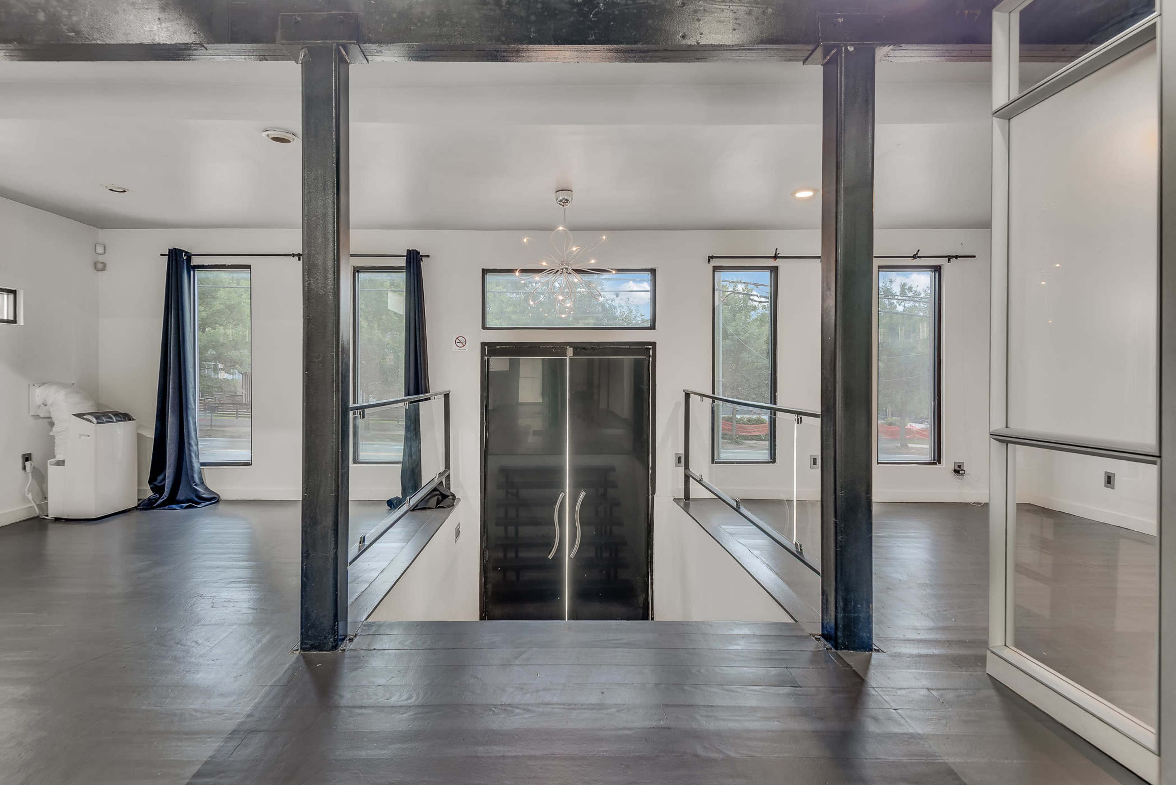 The image shows a modern, open interior space with large windows, a staircase leading down to an elevator, and sleek black flooring.