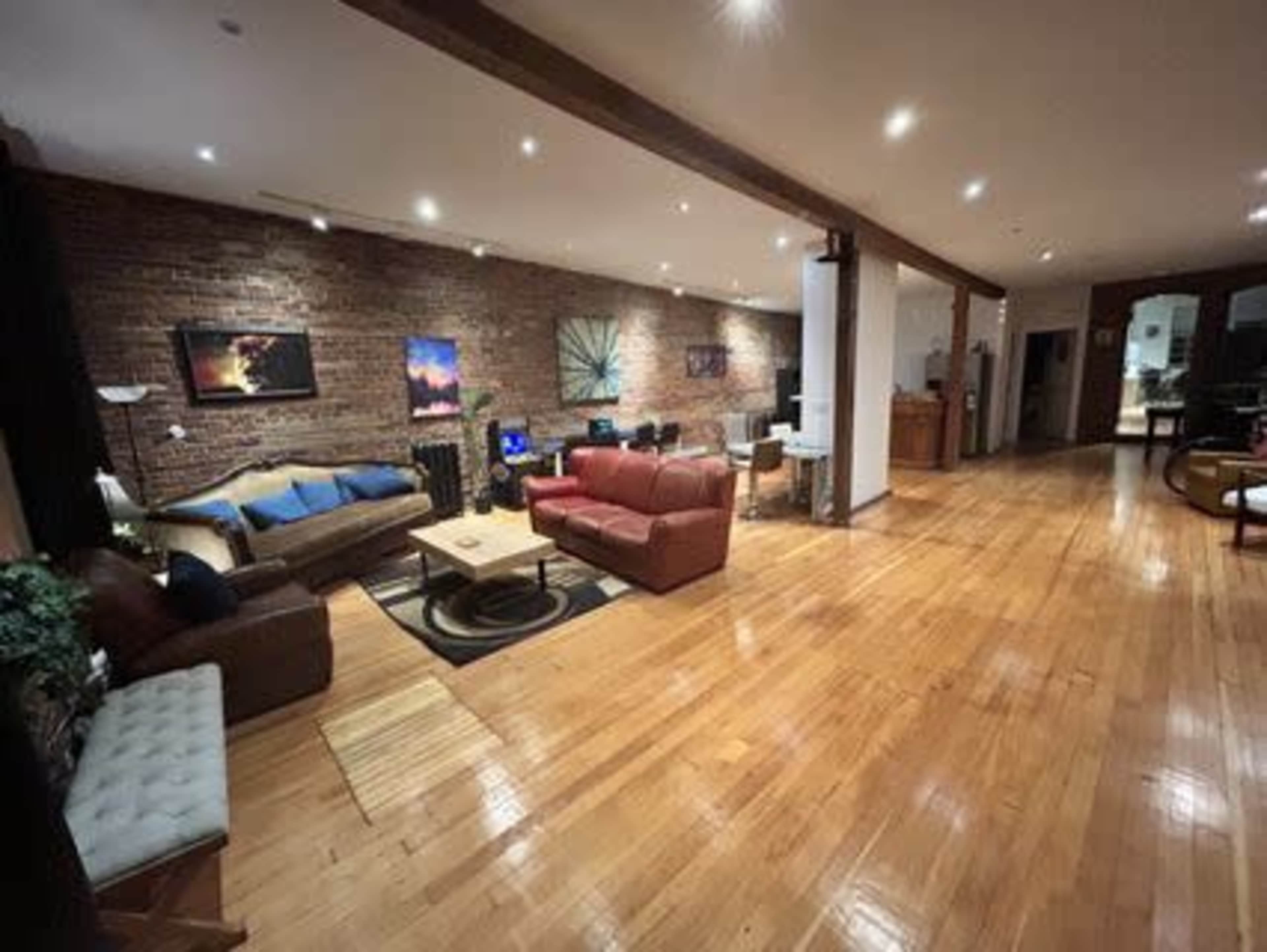 The image shows a spacious living area with wooden floors, featuring a mix of seating arrangements and a workspace area against a backdrop of exposed brick walls.