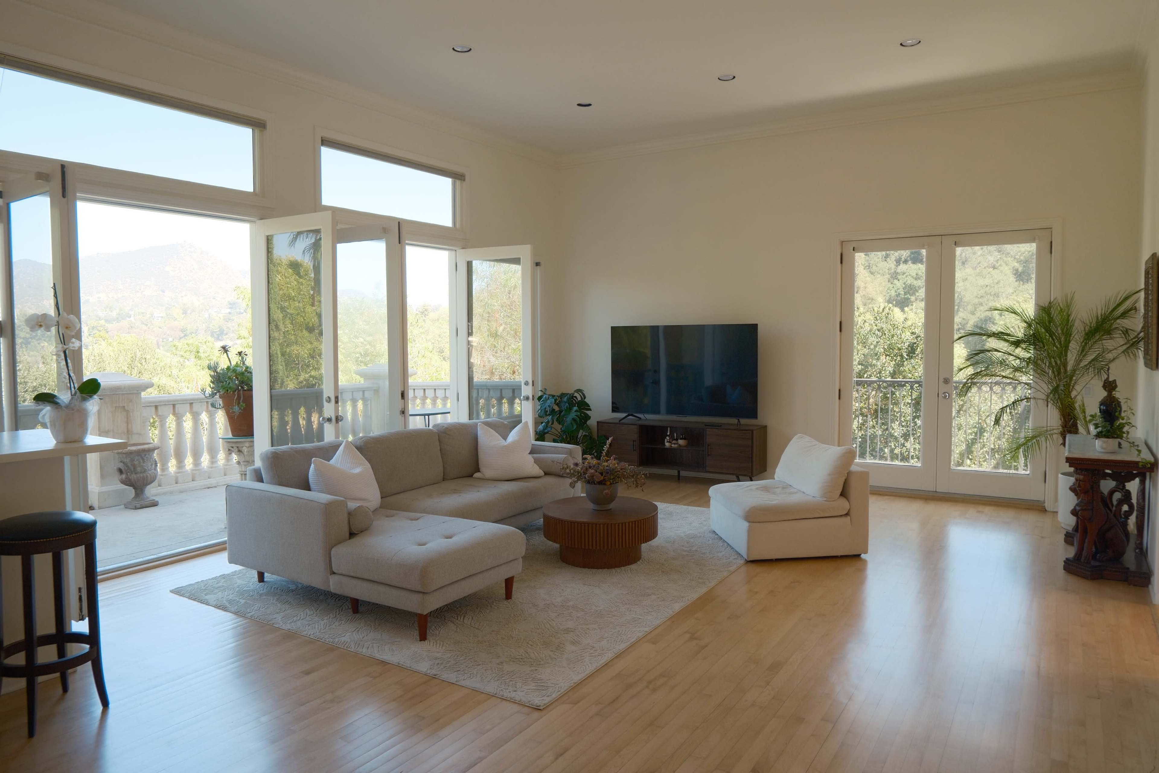 The image shows a spacious living room with large windows, a sectional sofa, a flat-screen TV, and plants, overlooking a scenic outdoor view.