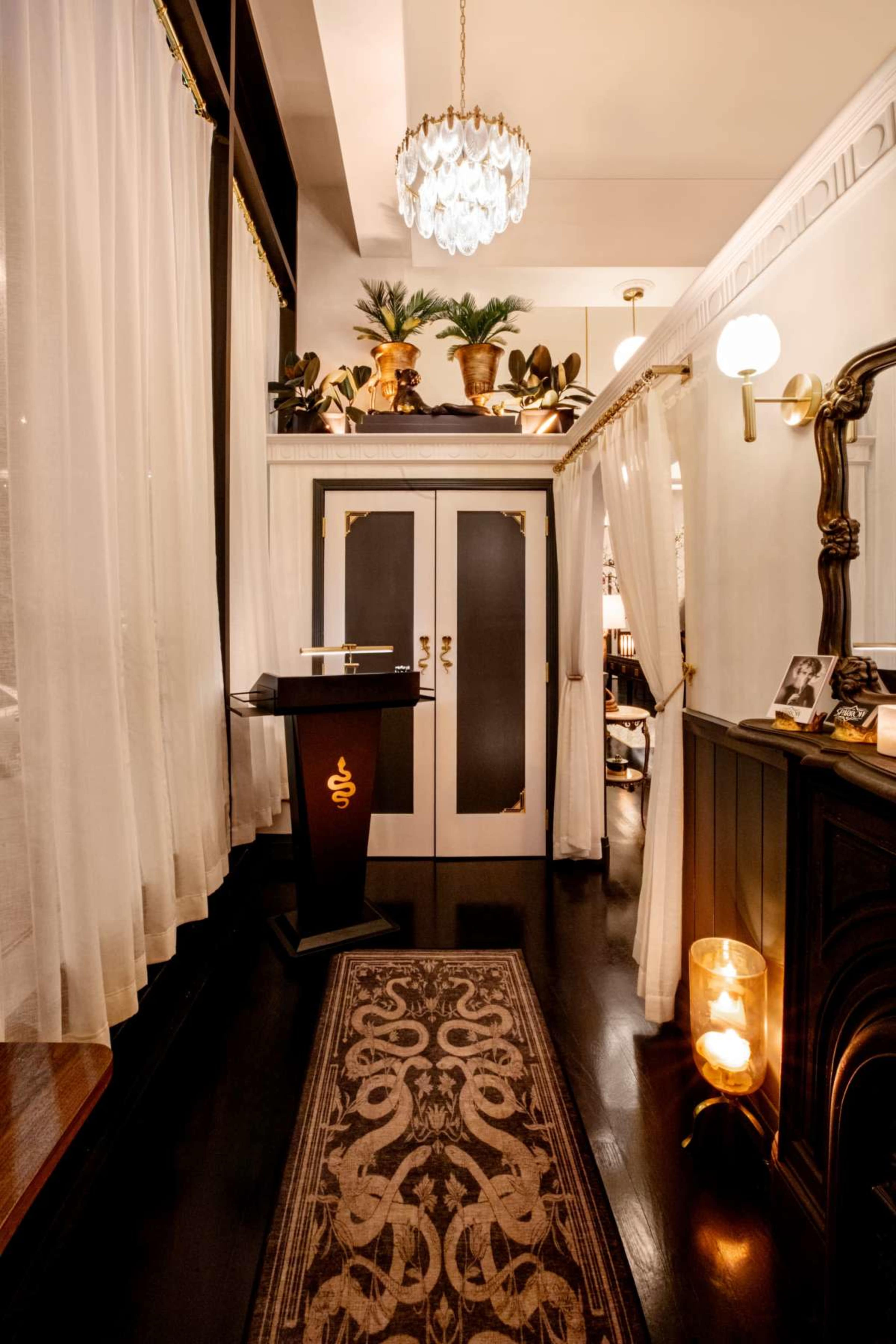 The image depicts a stylish interior hallway with sheer drapes, a decorative rug, and elegant lighting fixtures leading to double doors adorned with gold accents.