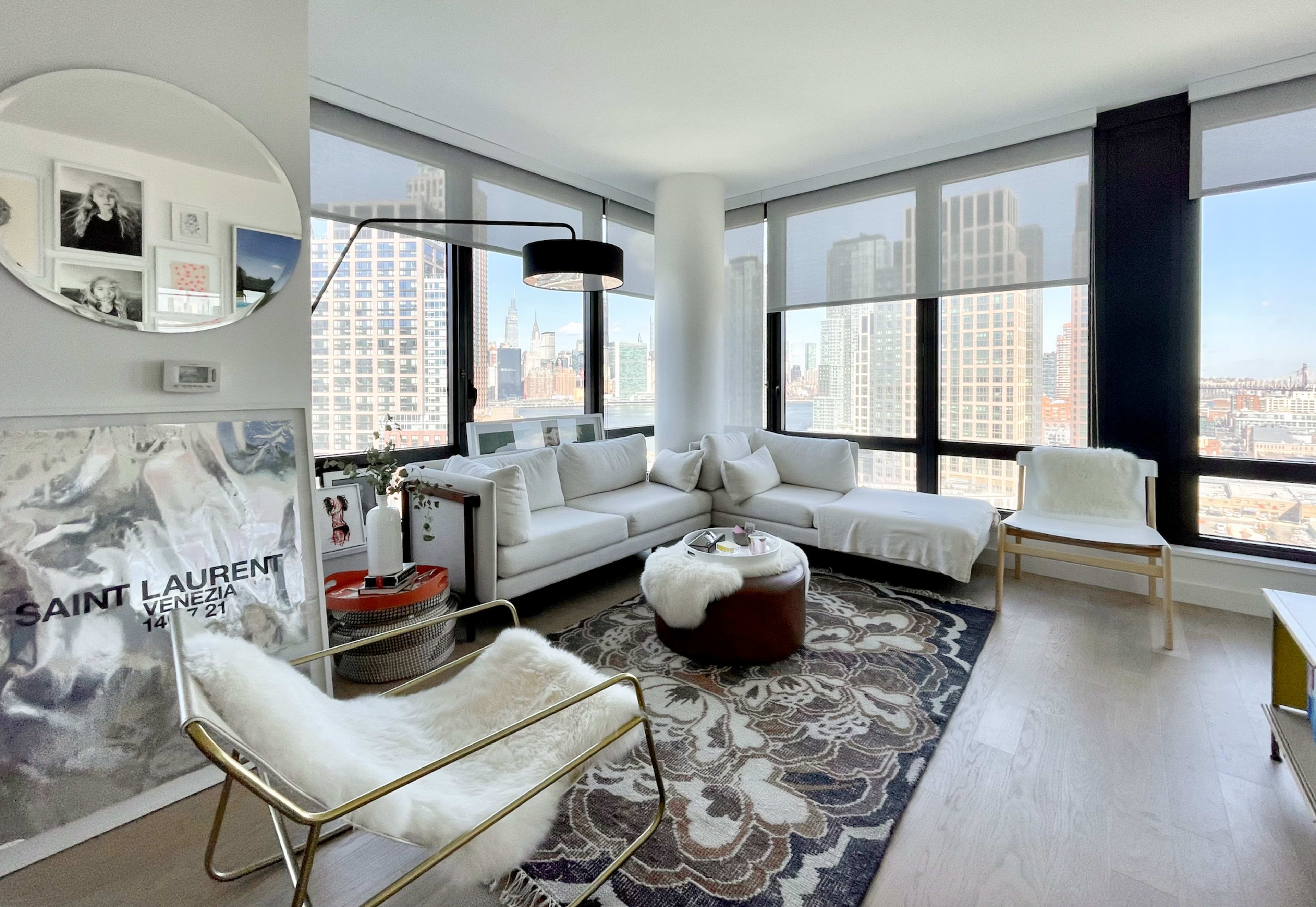 The image shows a modern living room with large windows overlooking a city skyline, featuring a white sectional sofa, a circular mirror, and a patterned rug.
