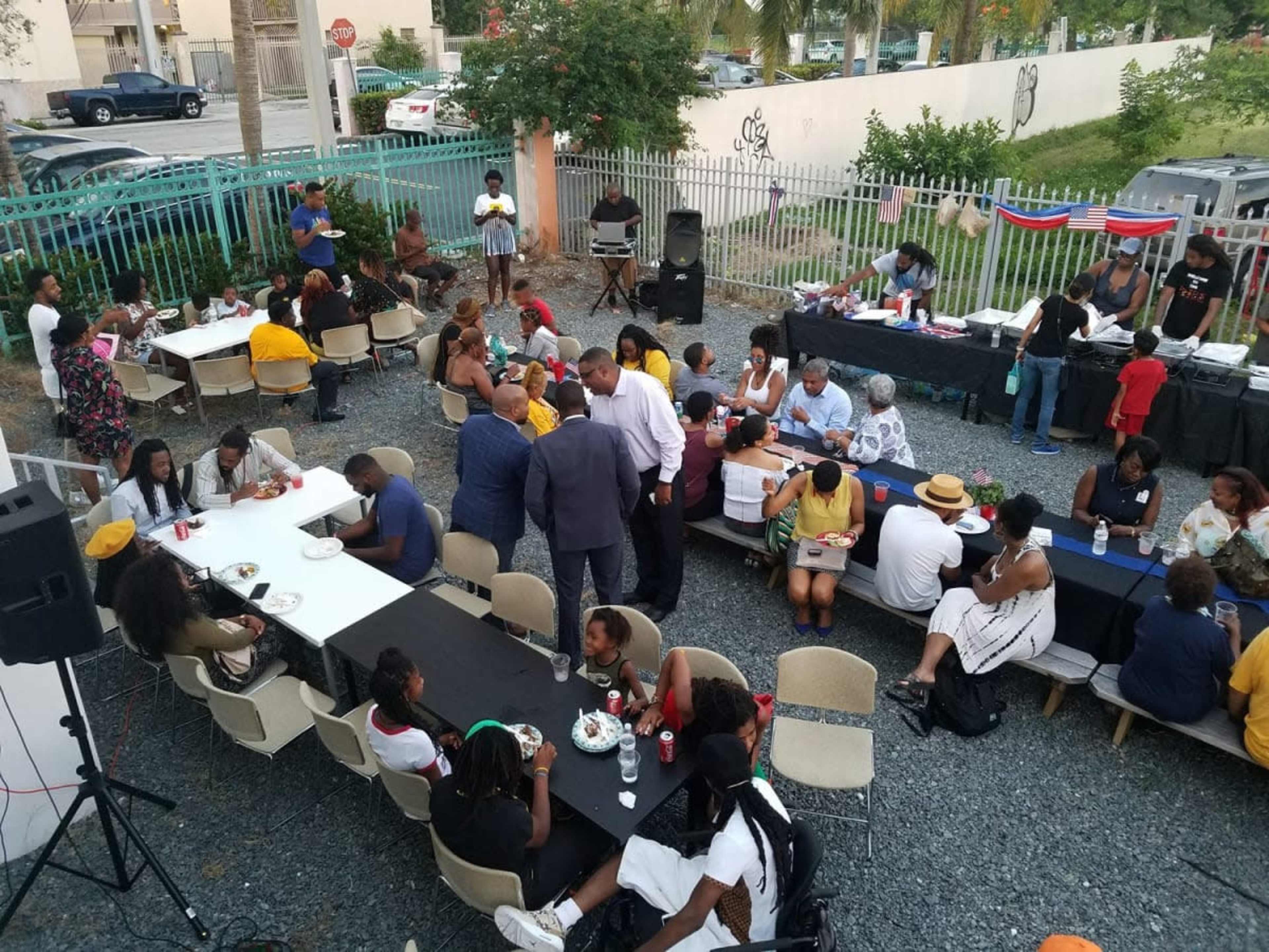 A large group of people gathers around several tables outdoors for a social event, with food and drinks served in a community setting.