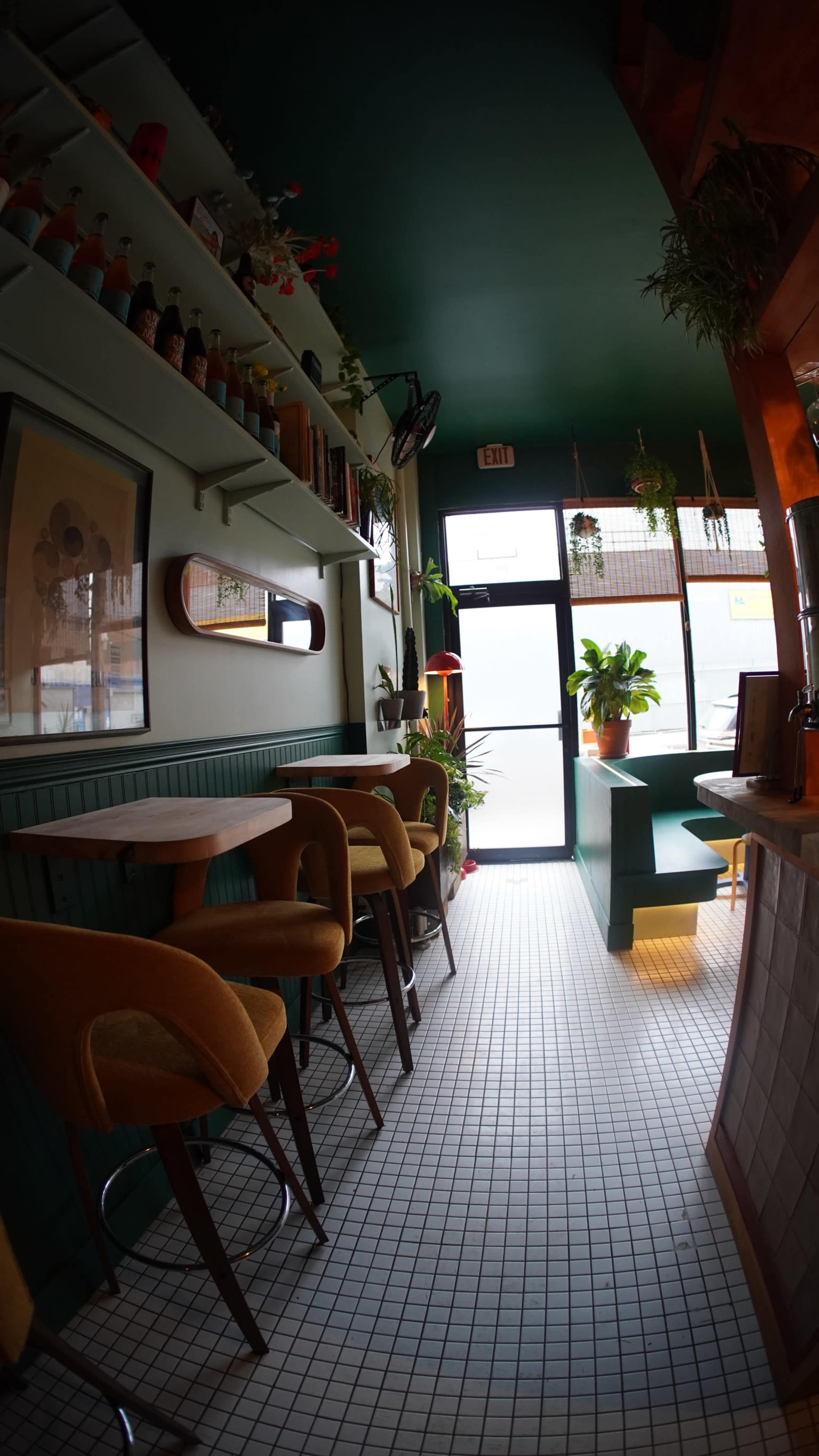 The image shows a cozy cafe interior featuring a green and white tiled floor, bar seating along the wall, and large windows letting in natural light.