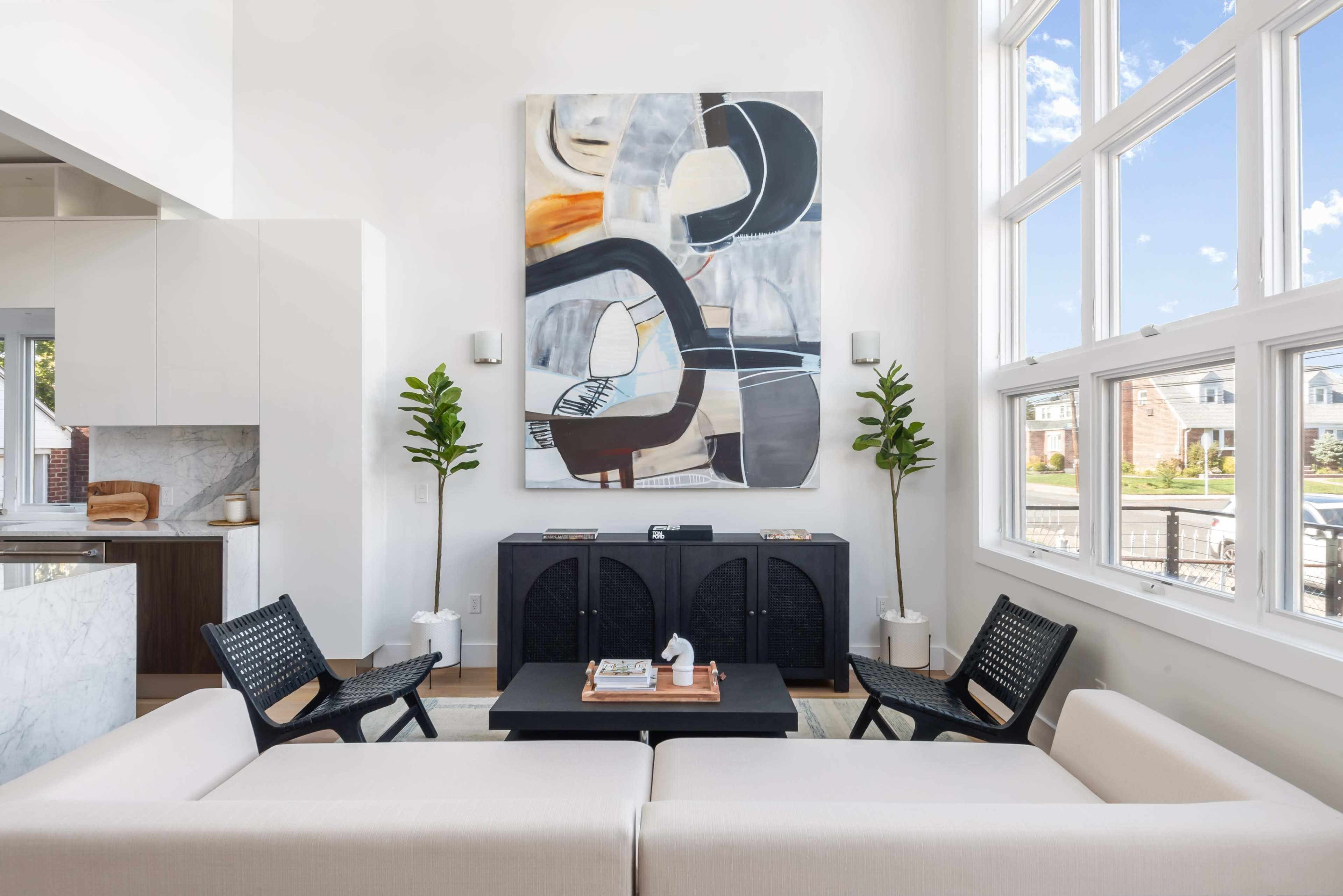 A modern living room features a large abstract painting above a minimalist black cabinet, flanked by two black chairs and a light-colored sofa.
