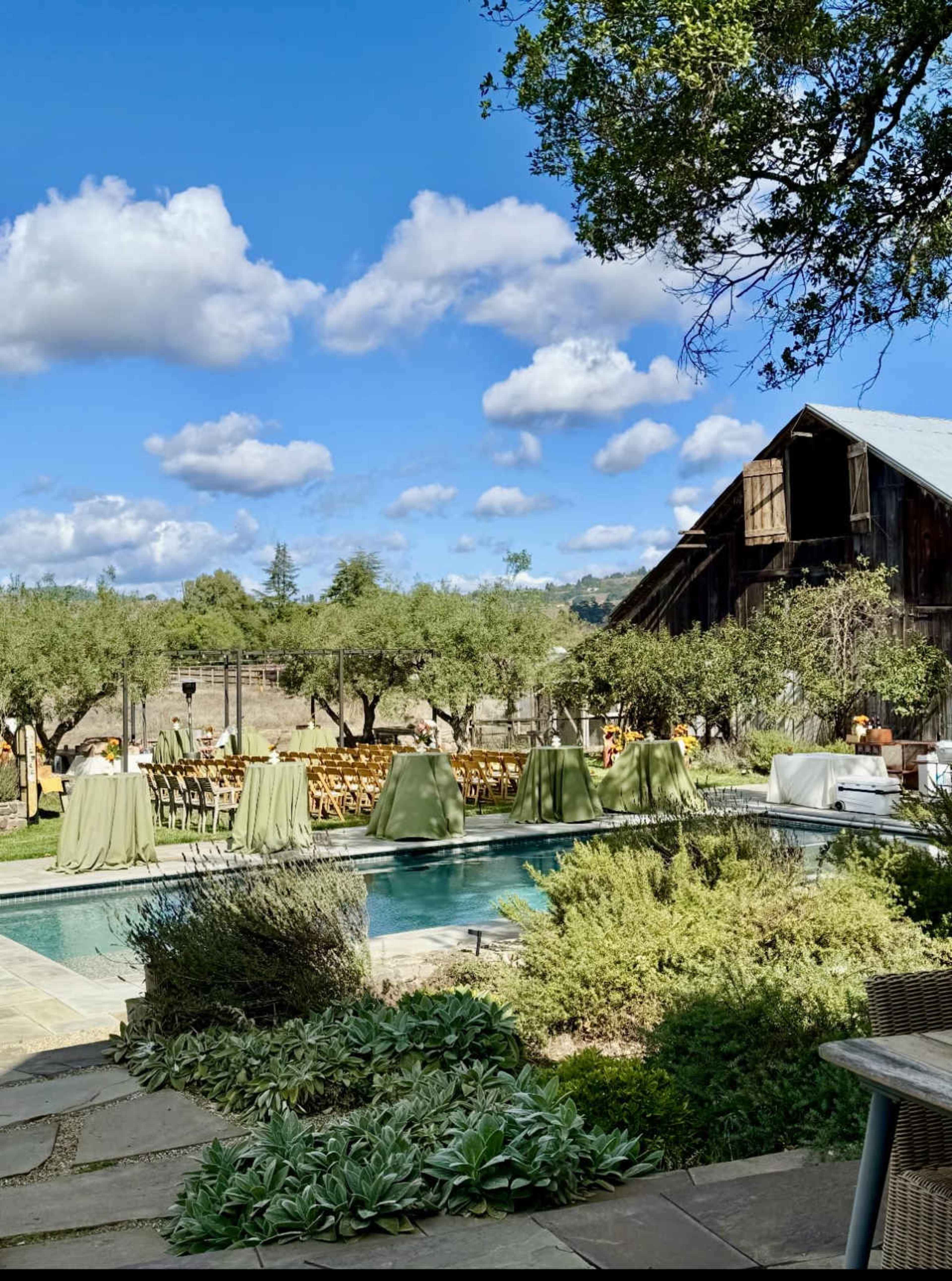 The image shows a swimming pool surrounded by greenery, with wooden chairs set up for an outdoor event near a rustic barn under a blue sky with scattered clouds.