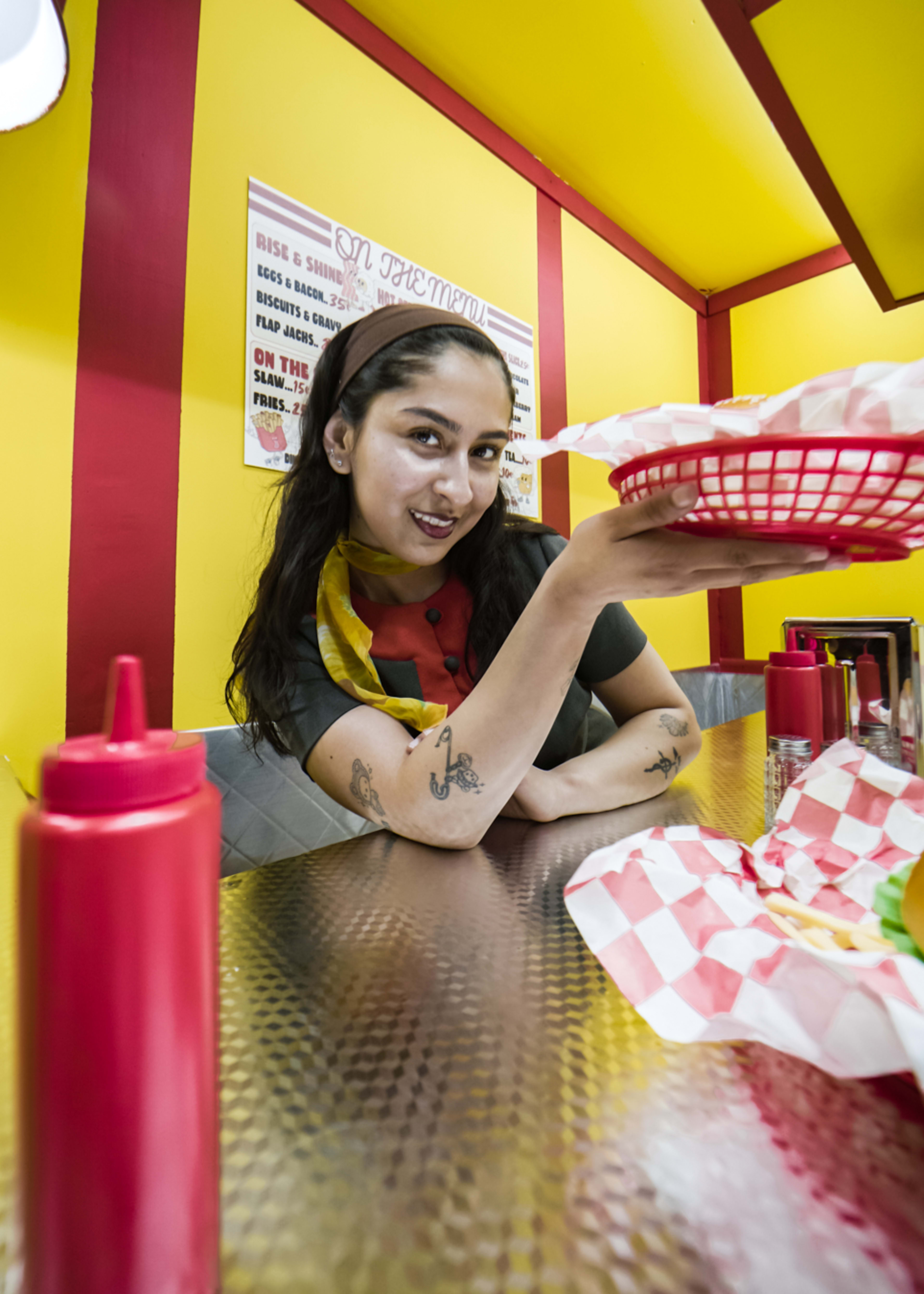 A restaurant employee smiles while holding a food basket in a brightly colored diner with red and yellow decor.