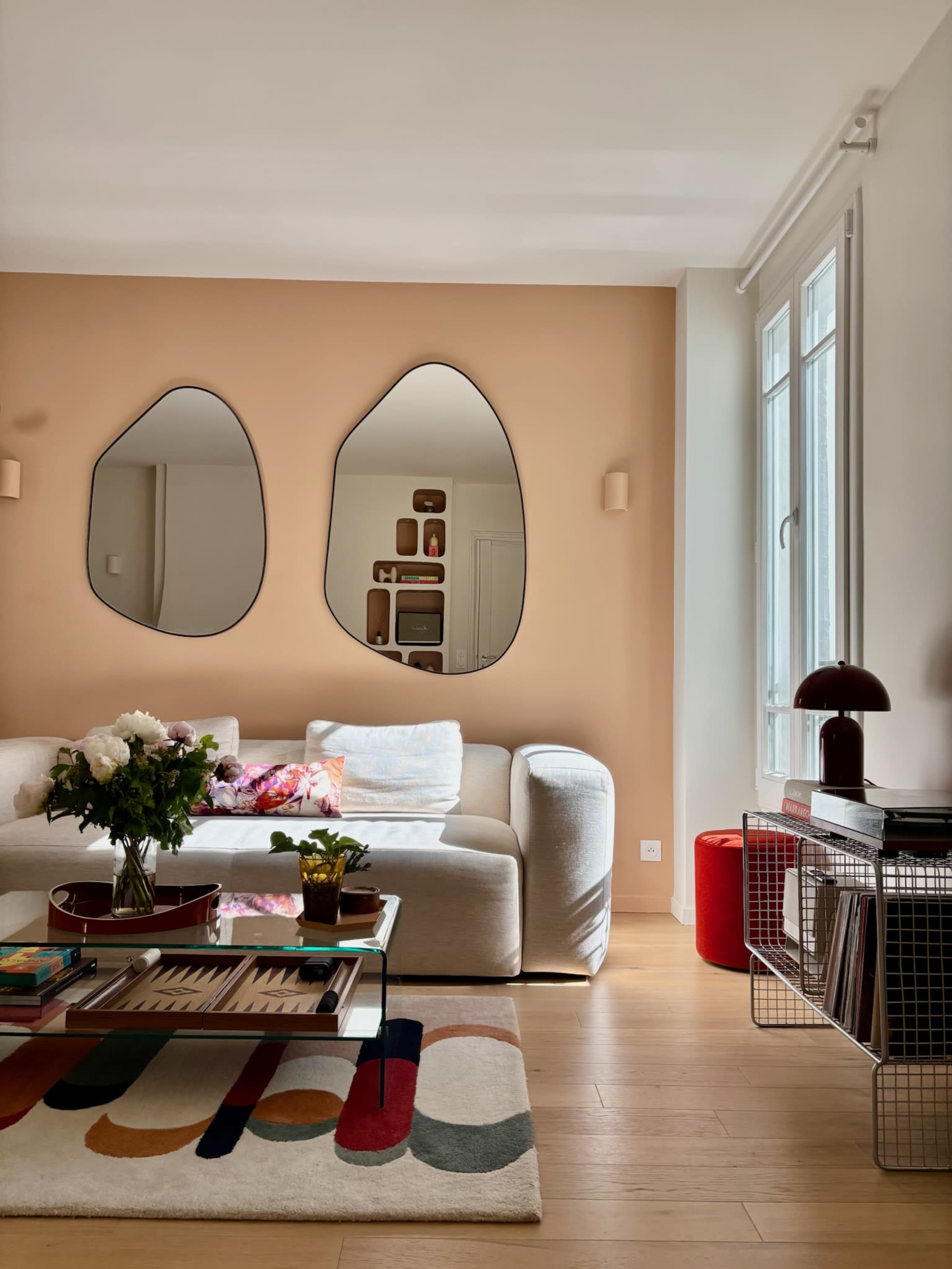 The image shows a modern living room with a light-colored sofa, a coffee table adorned with books and decor, and two abstract mirrors hanging on a peach-colored wall.