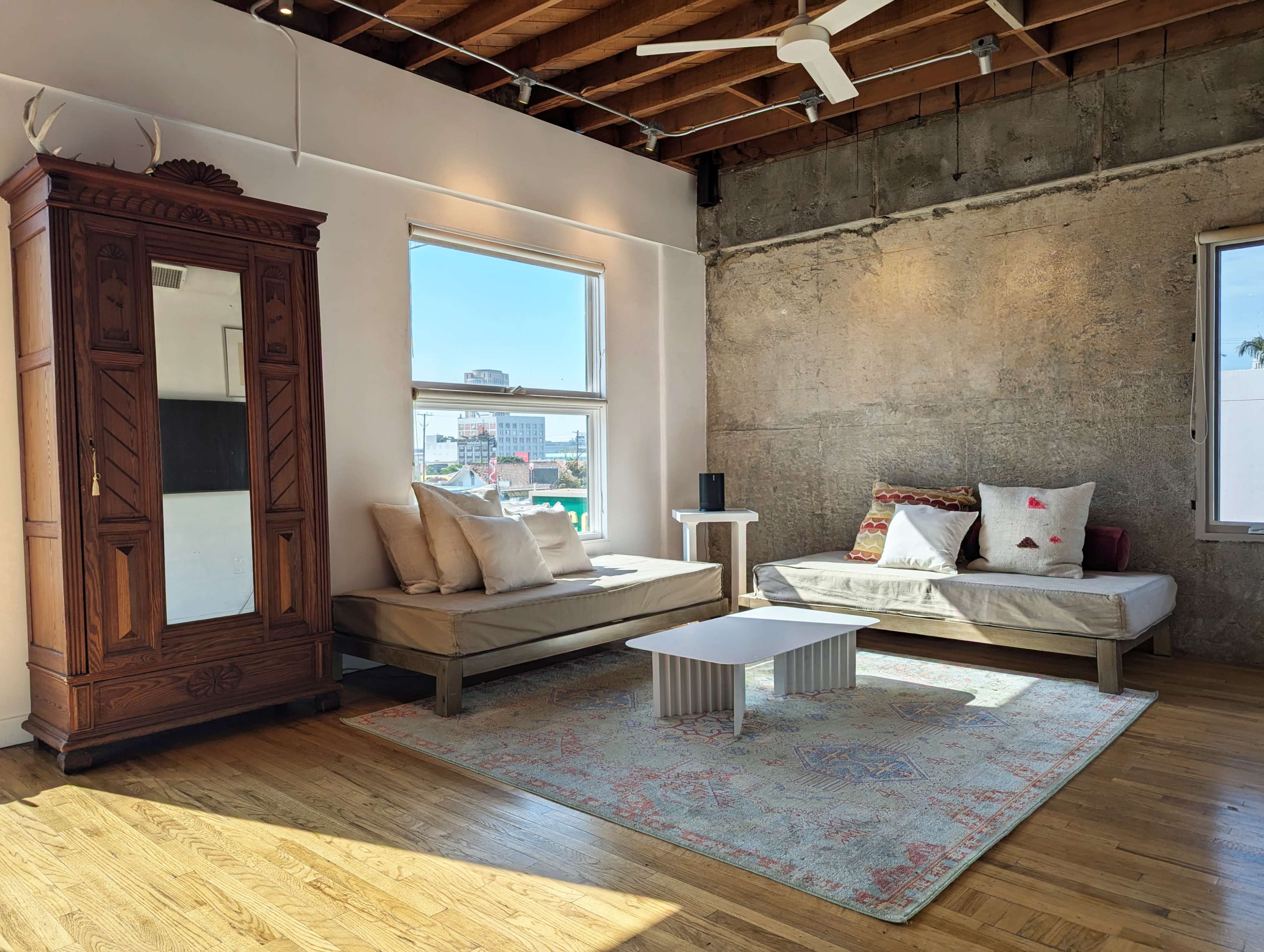 The image shows a bright living space featuring a wooden cabinet, two light-colored couches with decorative pillows, and a low white coffee table placed on a patterned rug.