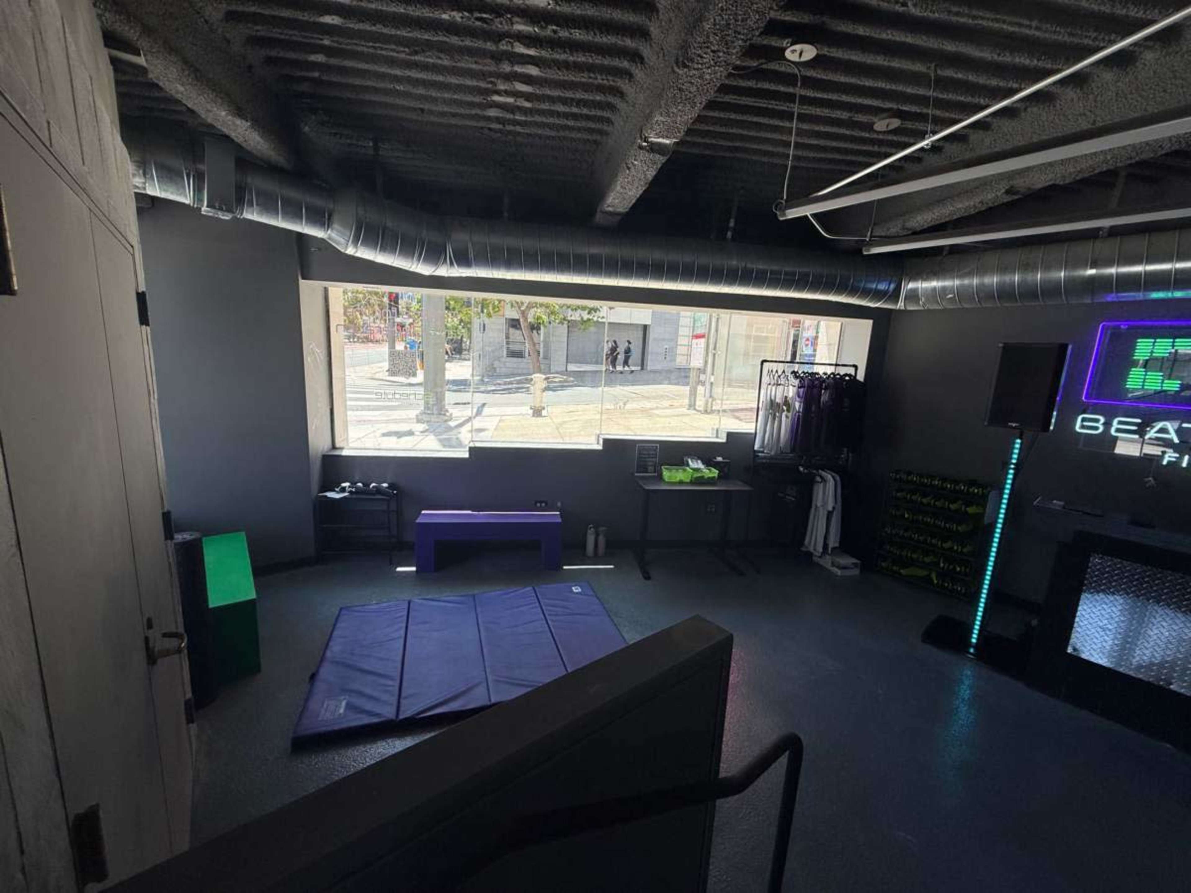 The image shows a gym studio with workout mats, exercise equipment, and large windows revealing a street view outside.