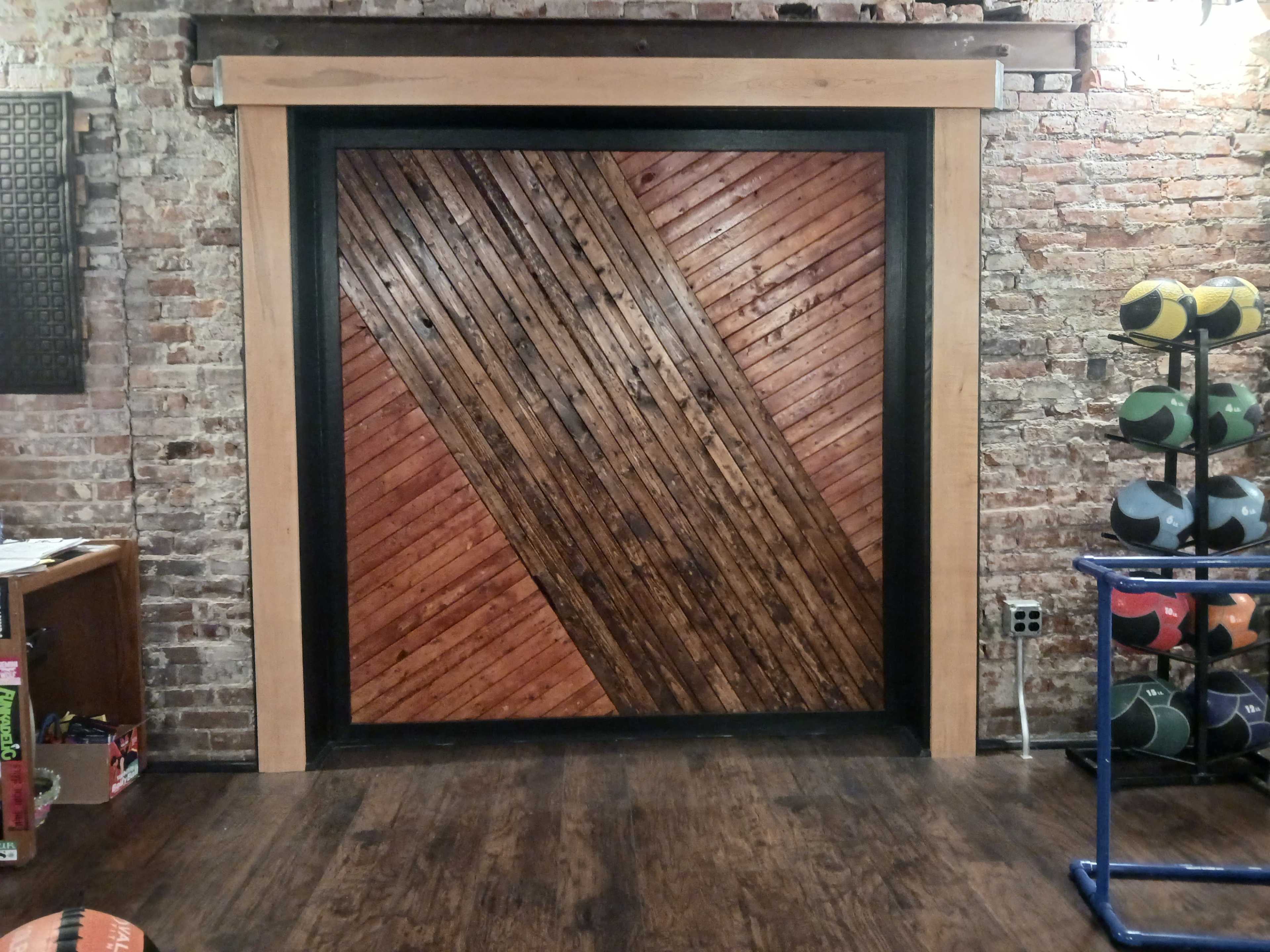 The image shows a wooden door with a diagonal patterned design, framed by black and natural wood trim, set against a backdrop of exposed brick walls.