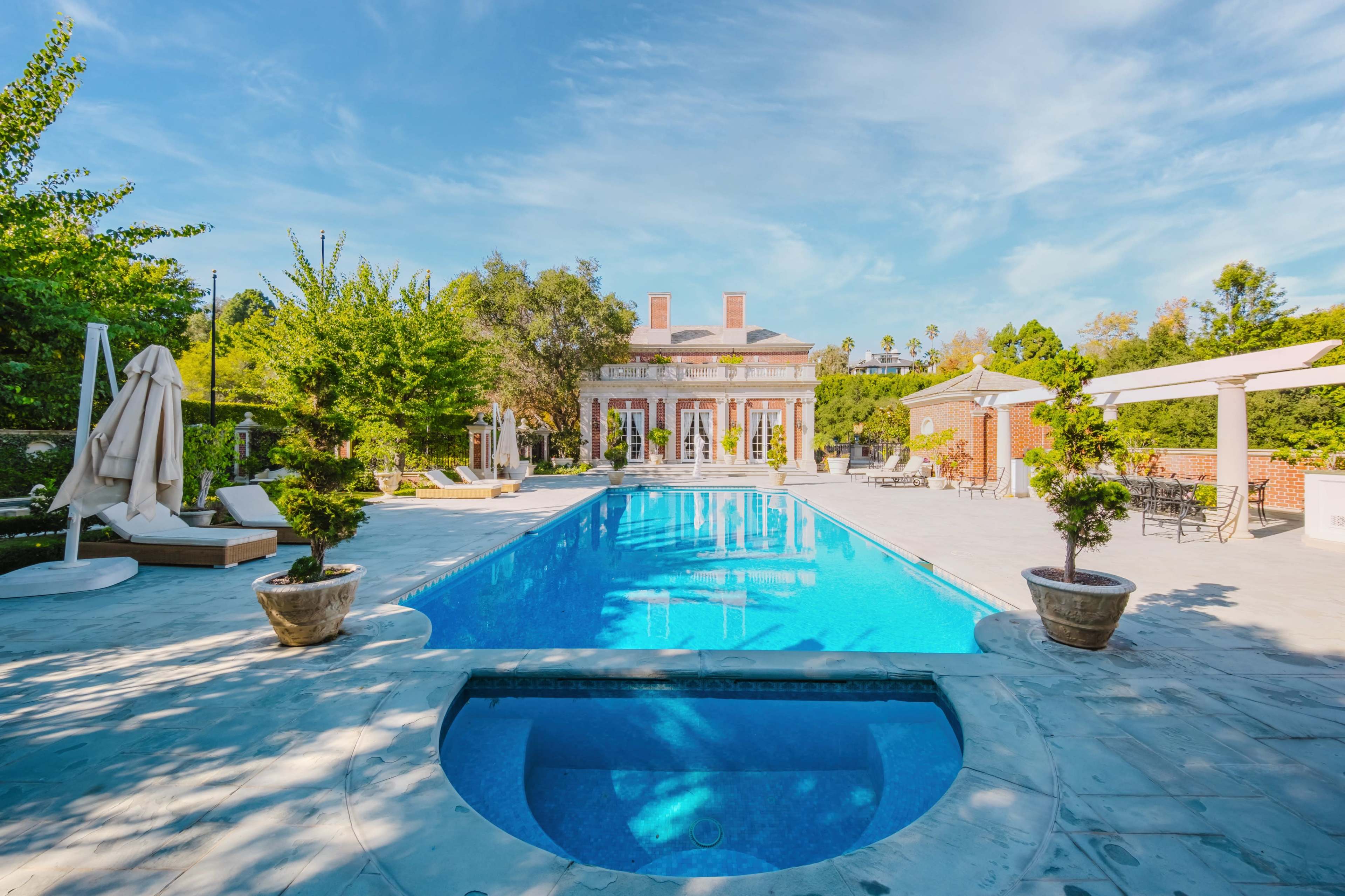 The image shows a spacious outdoor area featuring a swimming pool, lounge chairs, and a large house in the background surrounded by greenery.