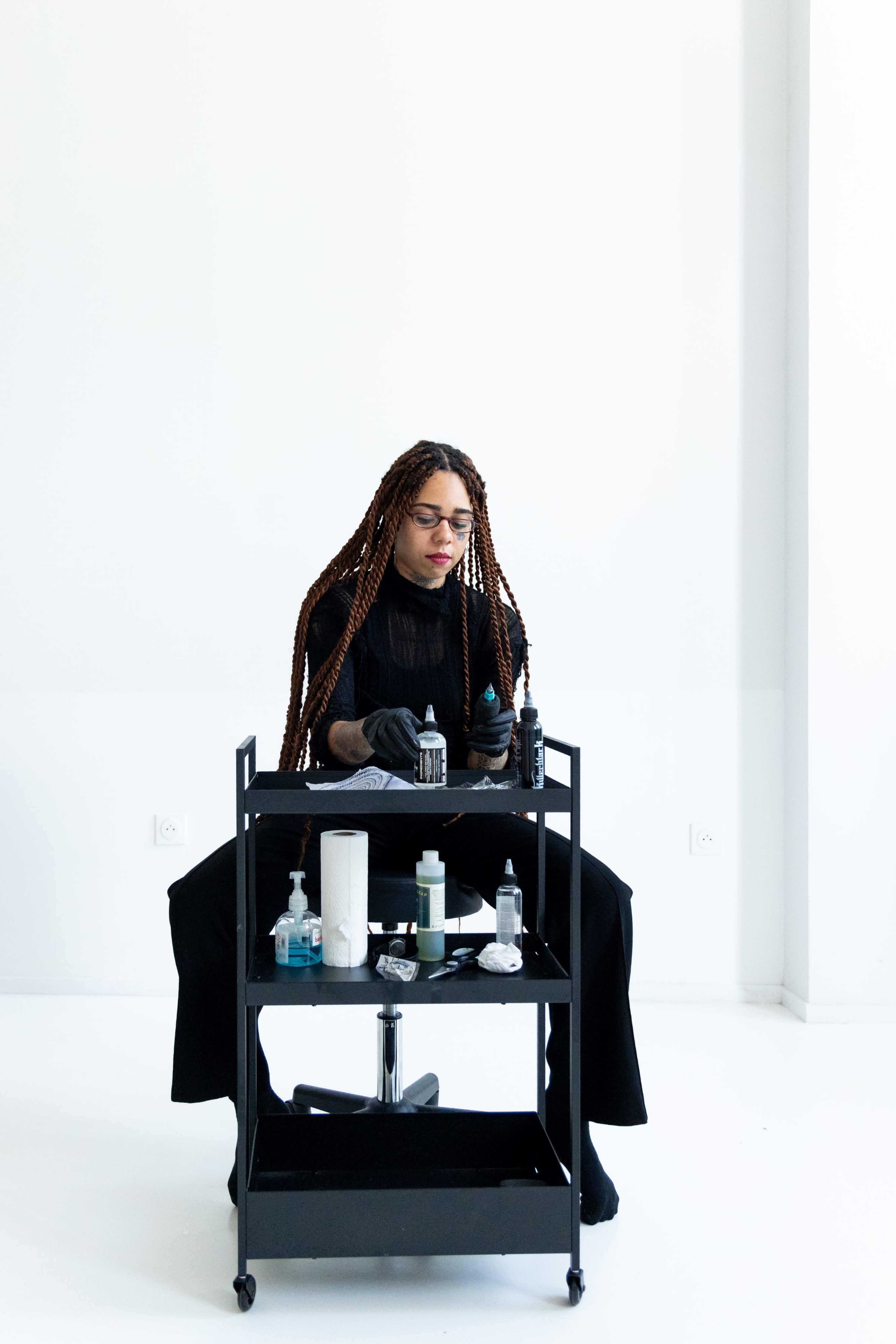 A person with long braided hair sits at a black workstation filled with various hair care products and tools in a minimalist white room.