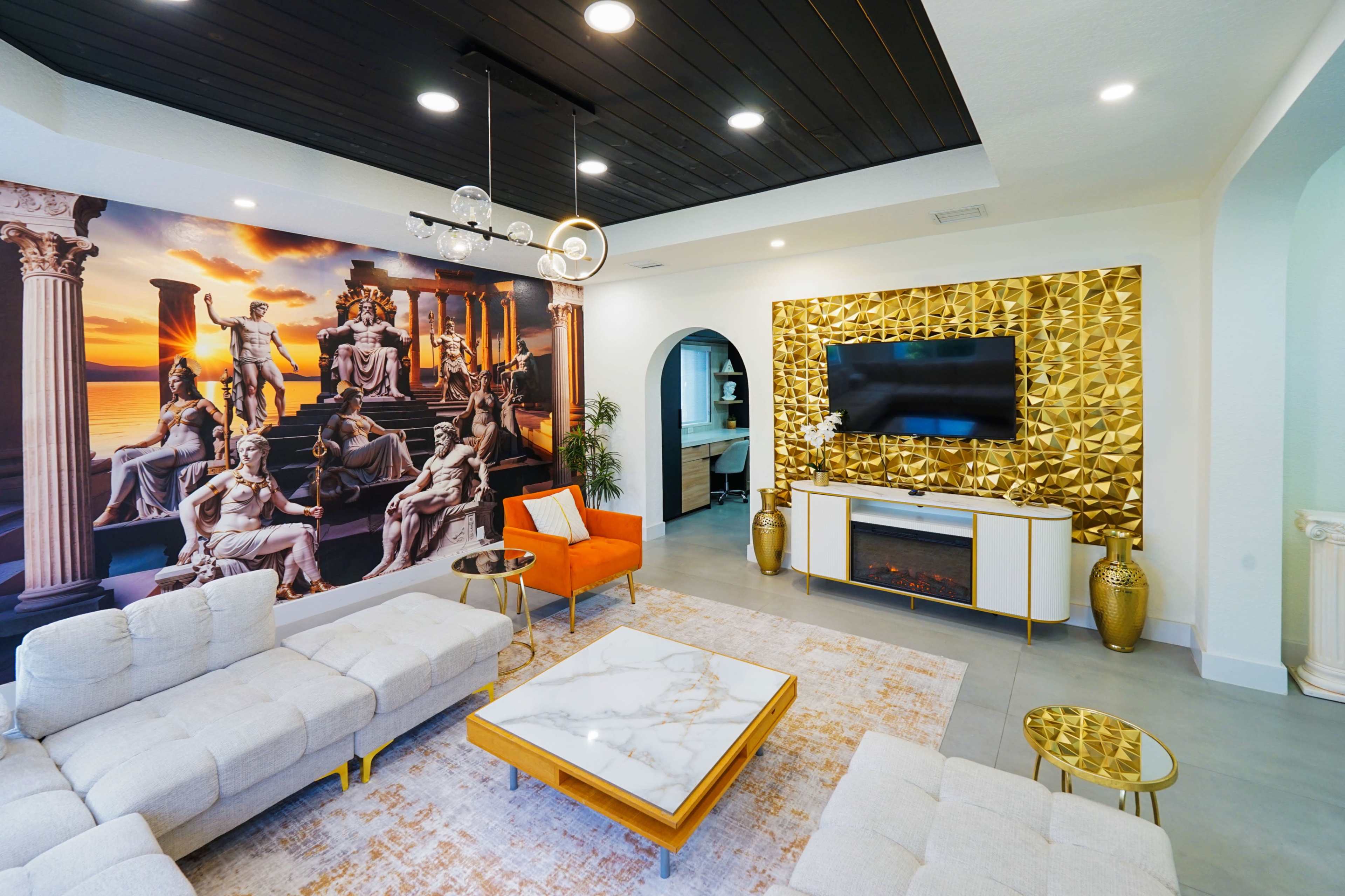 The living room features a modern design with a large mural of classical sculptures, a golden wall unit with a television, and contemporary furniture including a white sectional sofa and an orange accent chair.