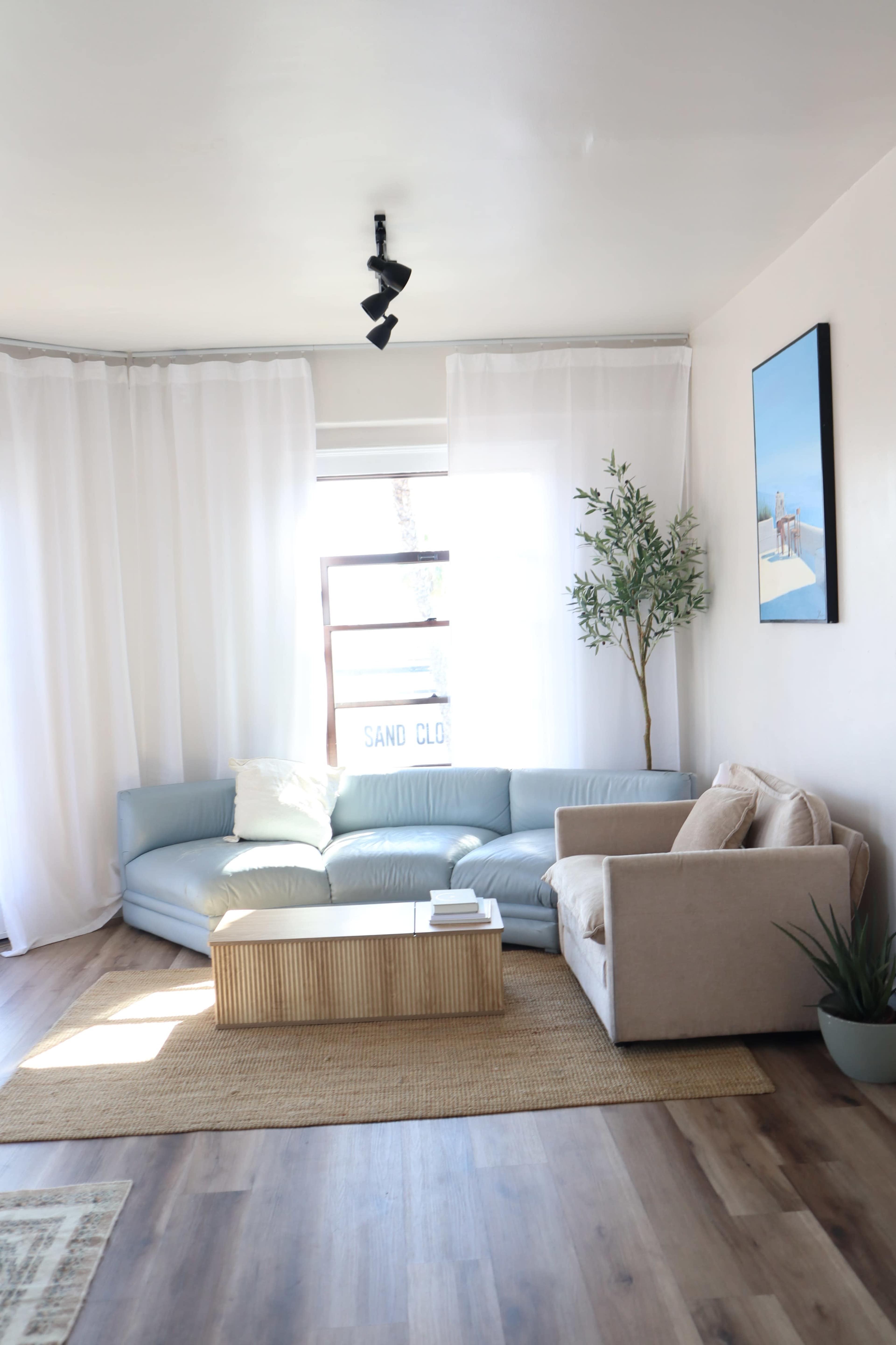 A modern living room features a curved blue sofa, a beige armchair, a wooden coffee table, and large windows covered by sheer white curtains.