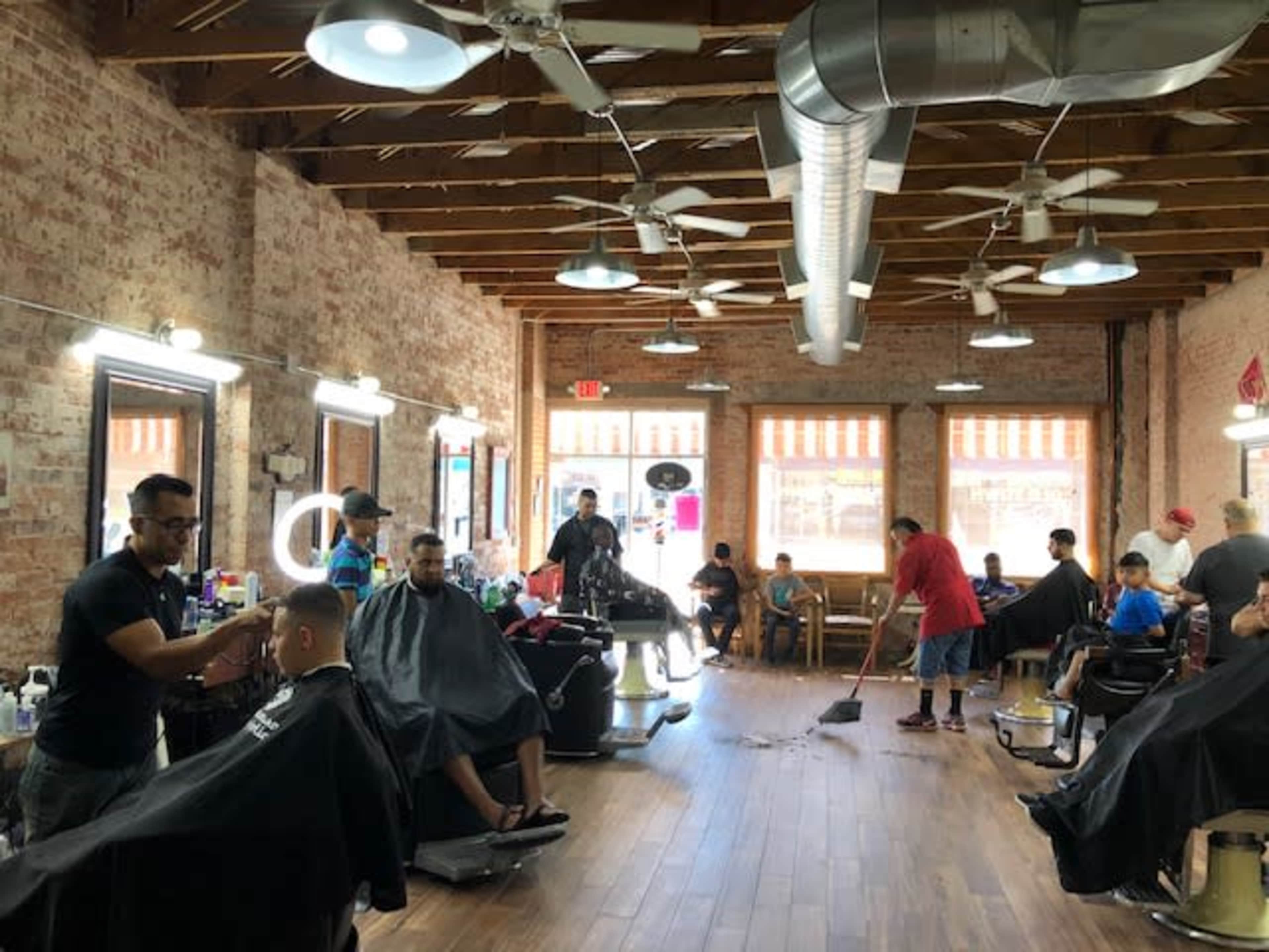 A busy barbershop features several customers in chairs, barbers working, and one person sweeping the floor.