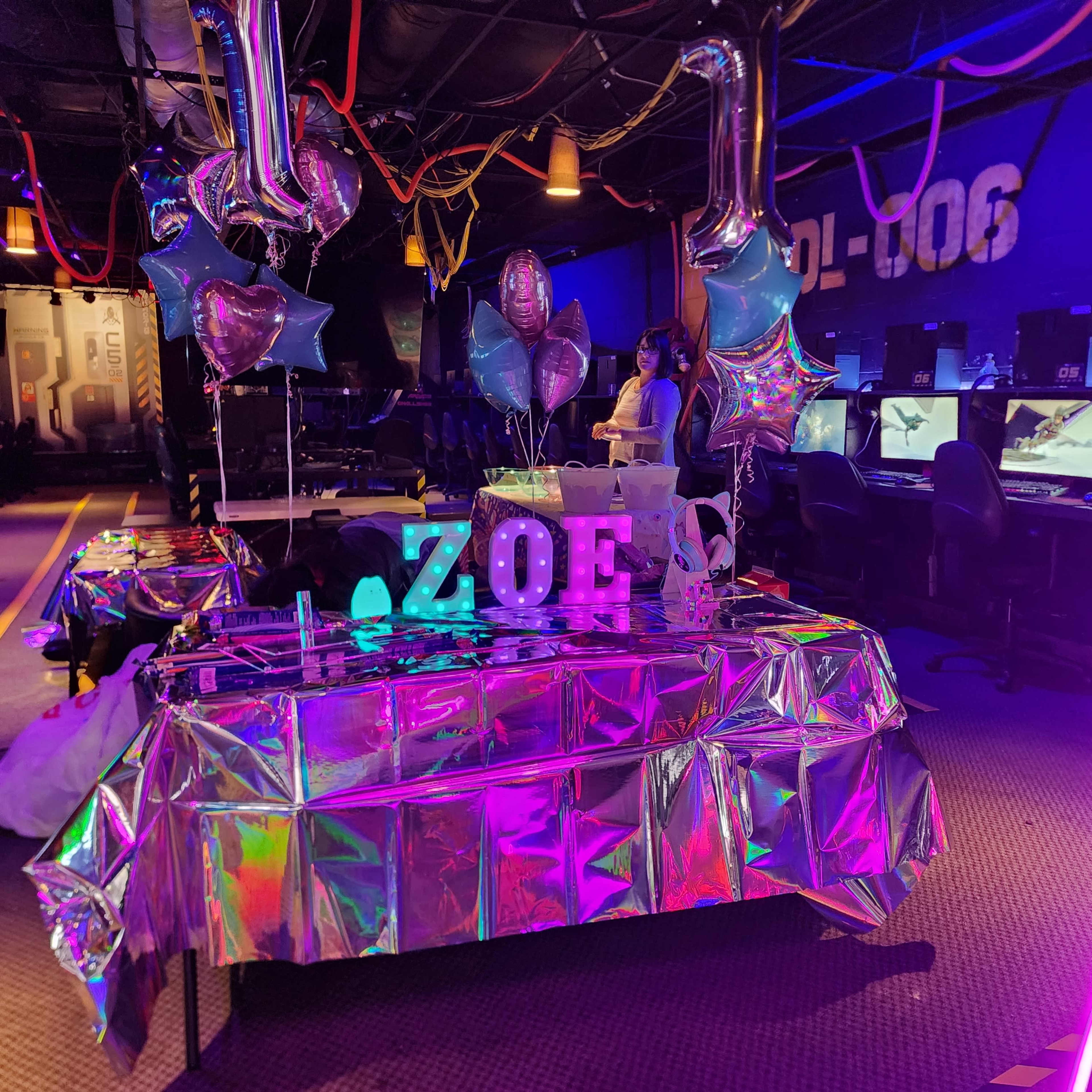 The image shows a decorated table with balloons and a sign that spells "ZOE" in a gaming arena setting.