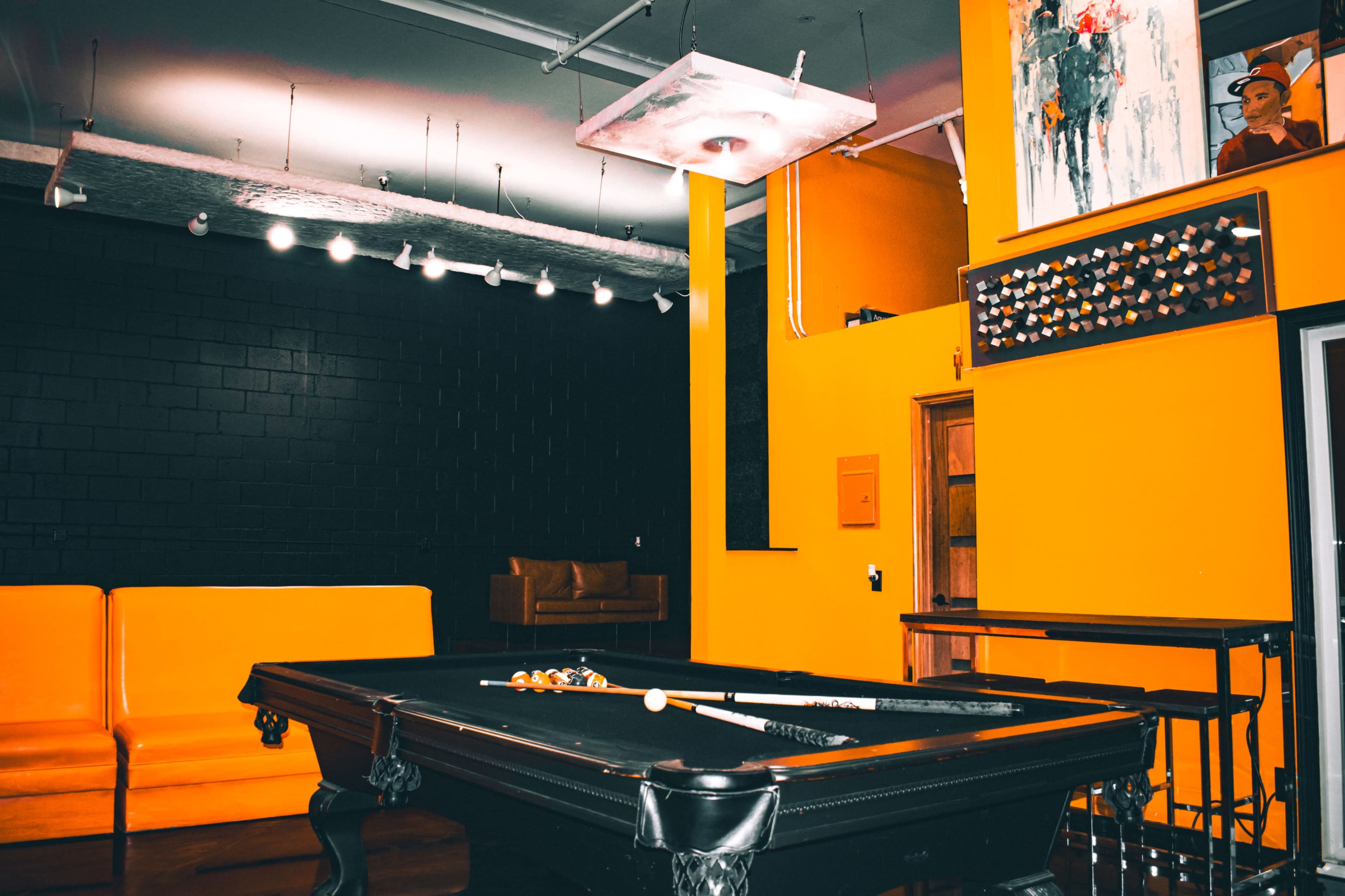 The image shows a brightly colored room featuring a black pool table, orange walls, and a seating area with a leather couch.