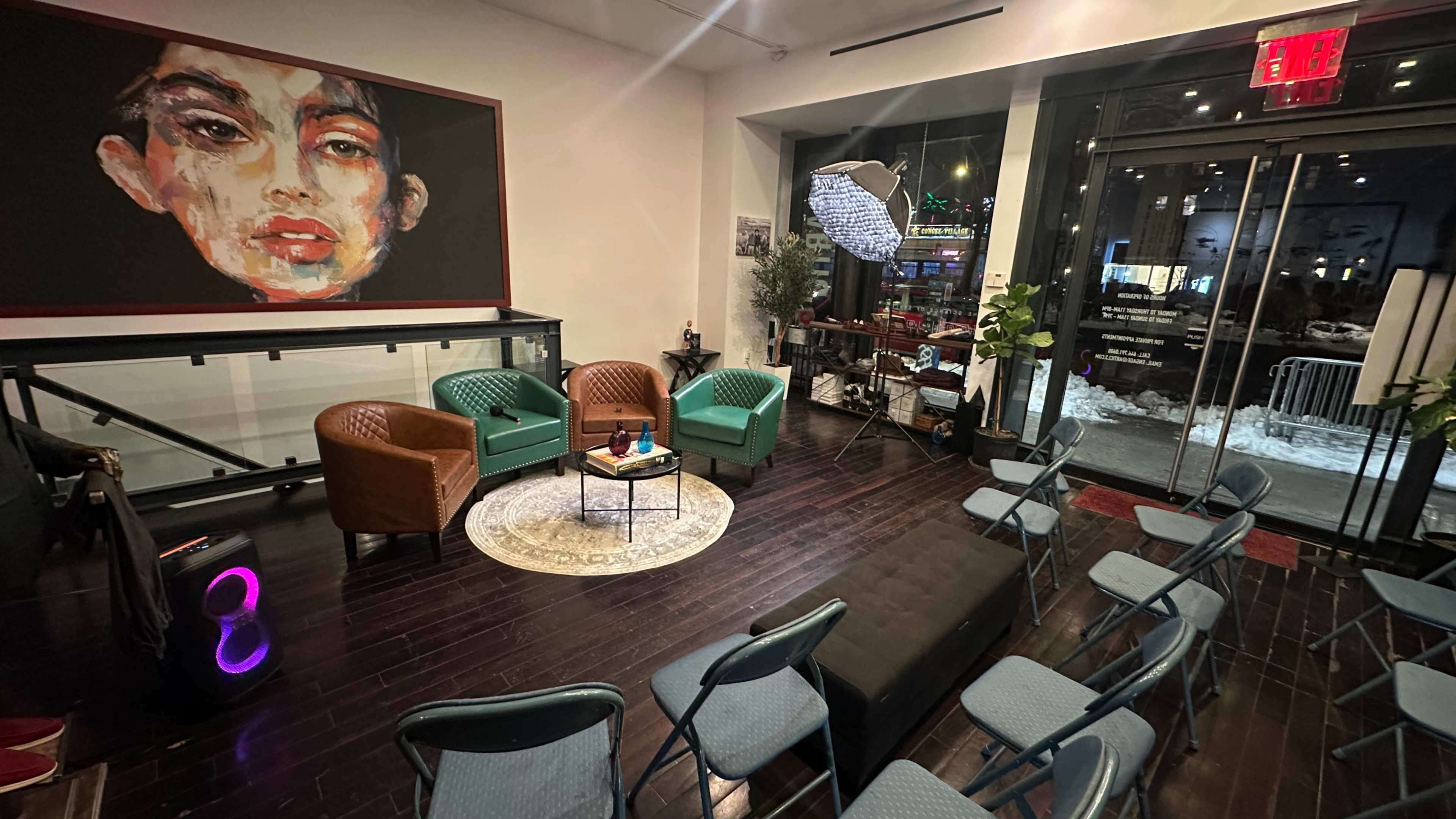 The image shows a modern indoor space with chairs arranged around a low table, green and brown seating, and a large colorful artwork on the wall.