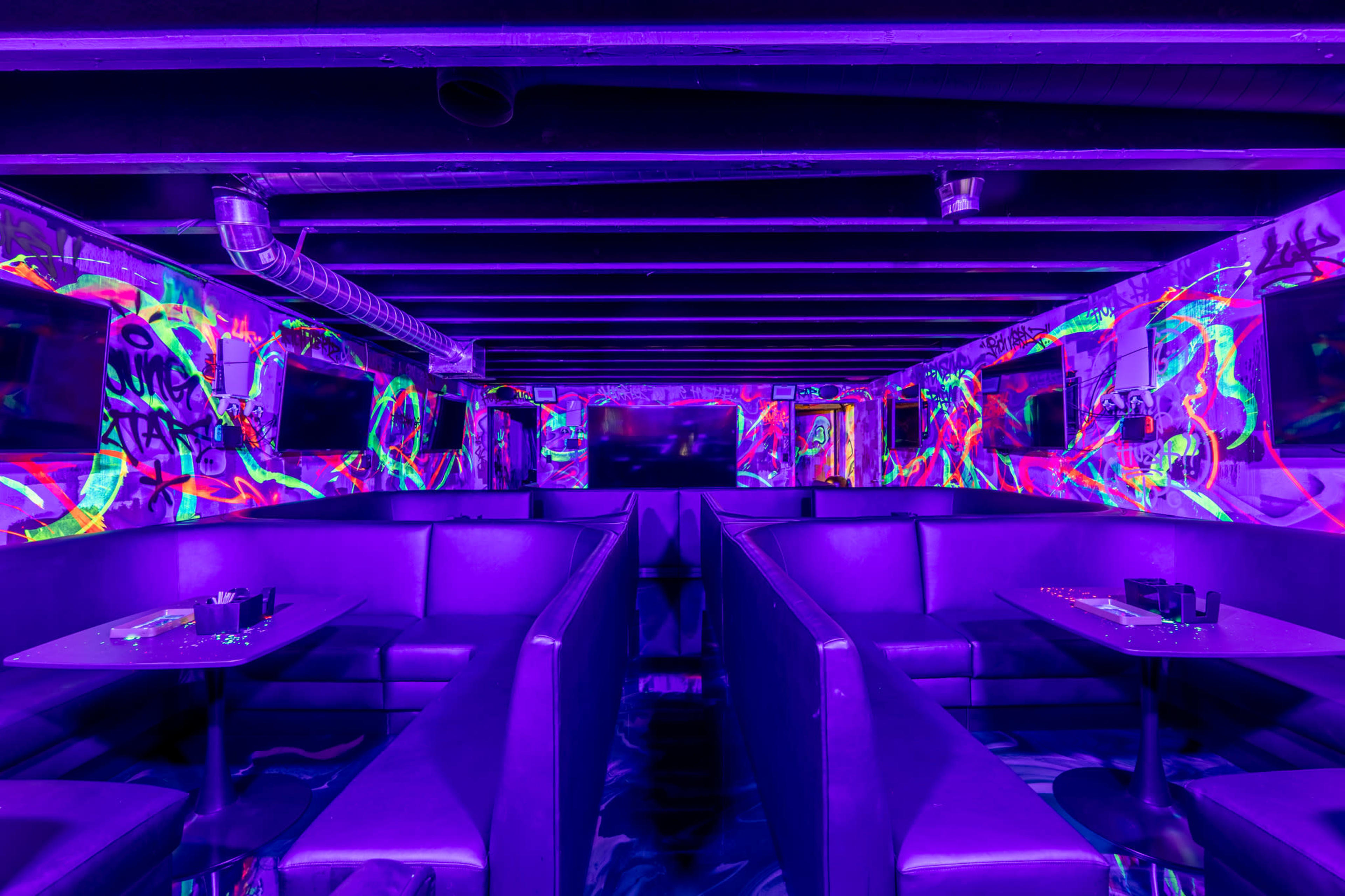The interior of a lounge is filled with vibrant, multicolored graffiti and illuminated by purple lighting, featuring booth seating and modern decor.
