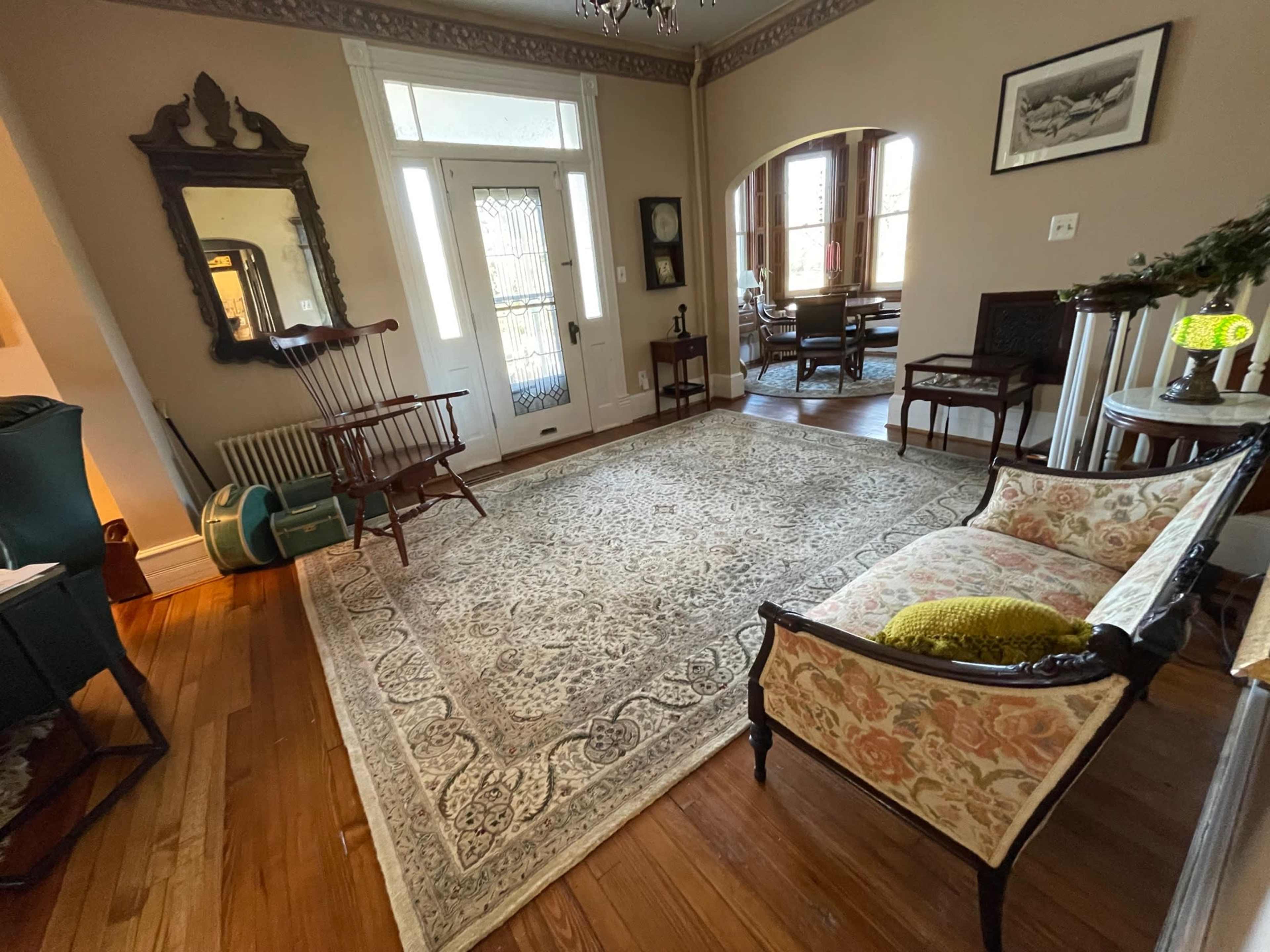 A spacious living room with wooden floors, a decorative rug, a vintage chair, and large windows allowing natural light.