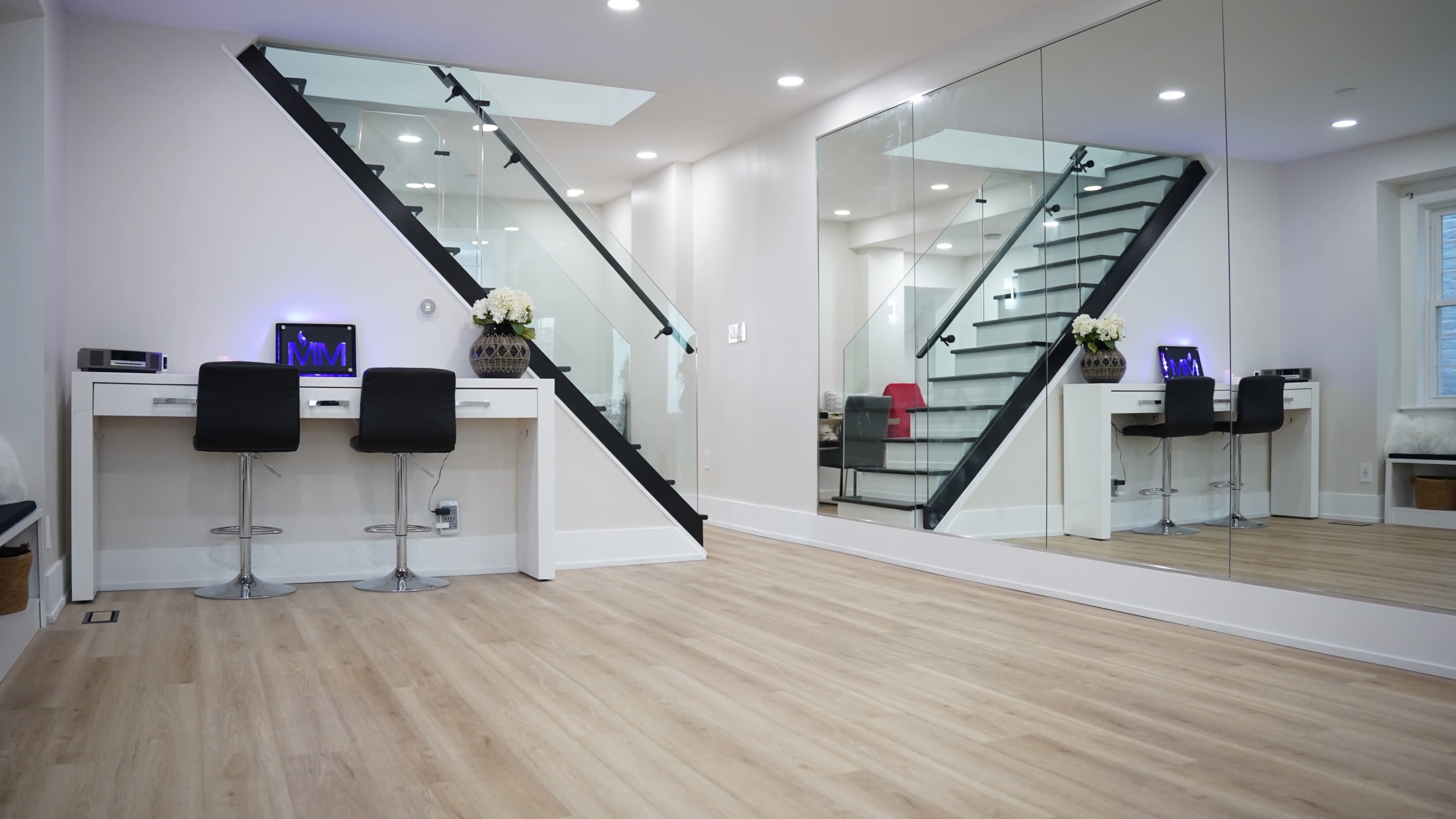The image shows a modern interior space featuring a staircase with glass panels, a desk with two black chairs, and large mirrors reflecting the surroundings.