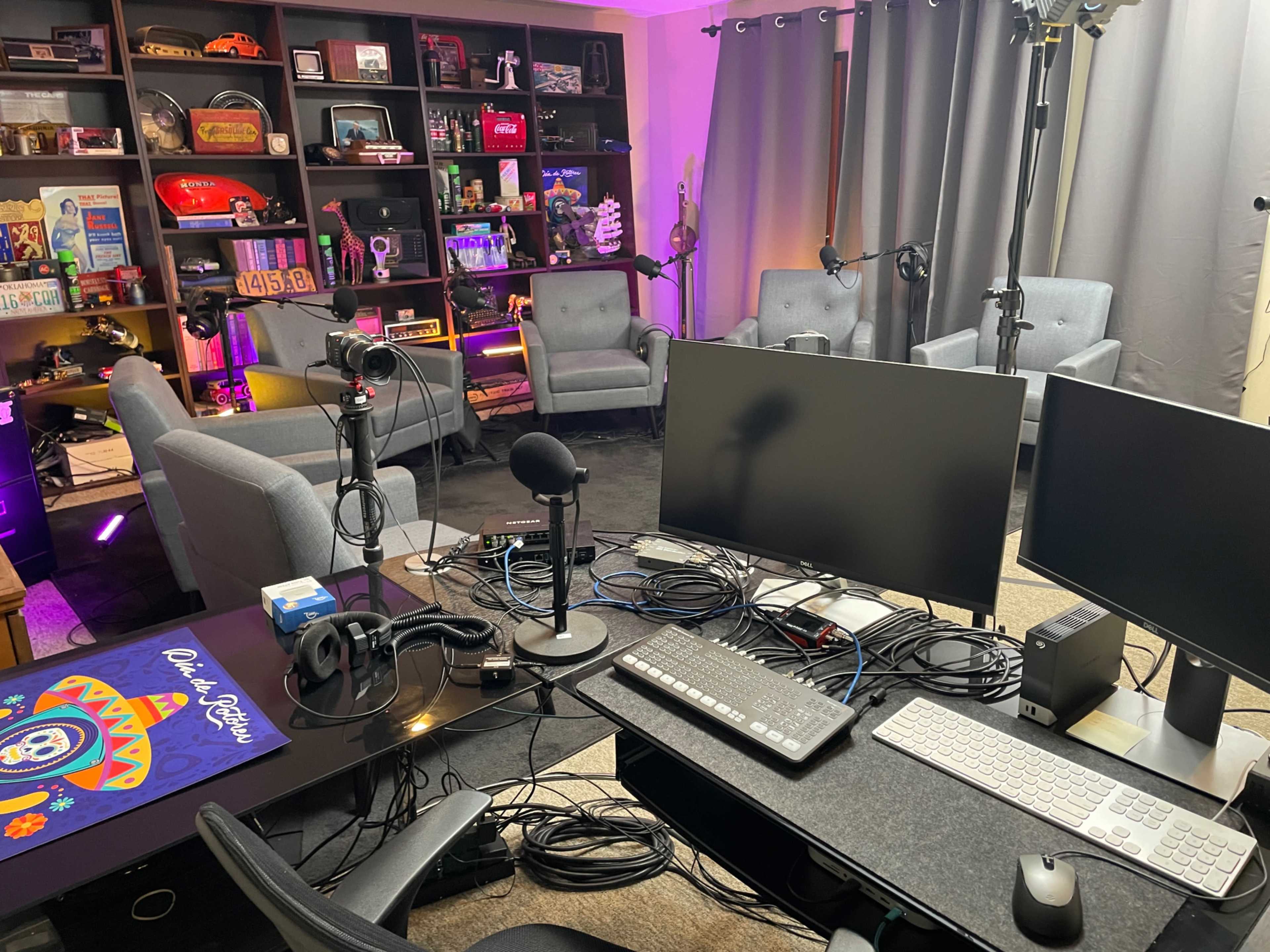 The image shows a modern audio-visual studio with multiple monitors, microphones, and a cluttered workspace, surrounded by seating arrangements and shelves filled with various items.
